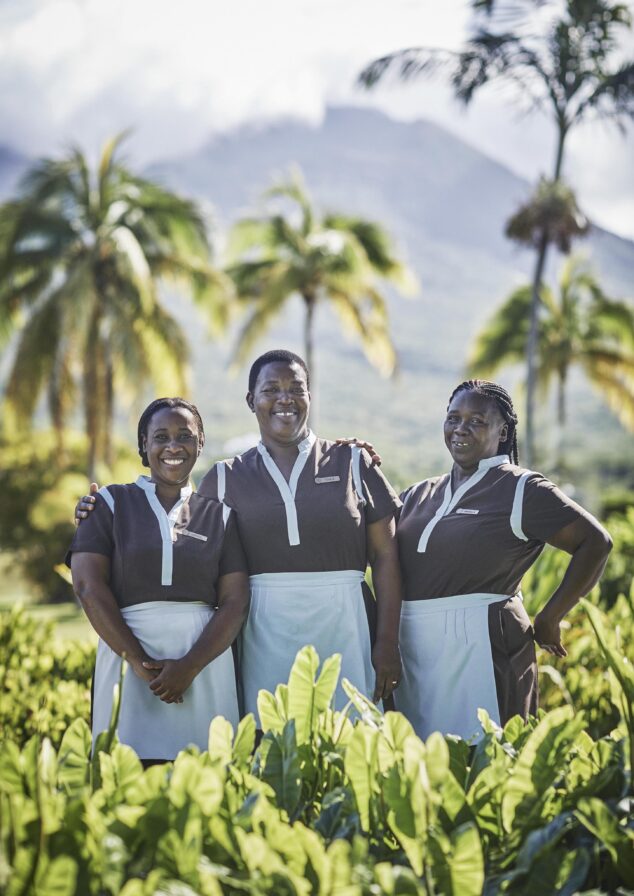 Three people in matching uniforms stand smiling in a lush garden with palm trees and a mountain in the background on a sunny day.
