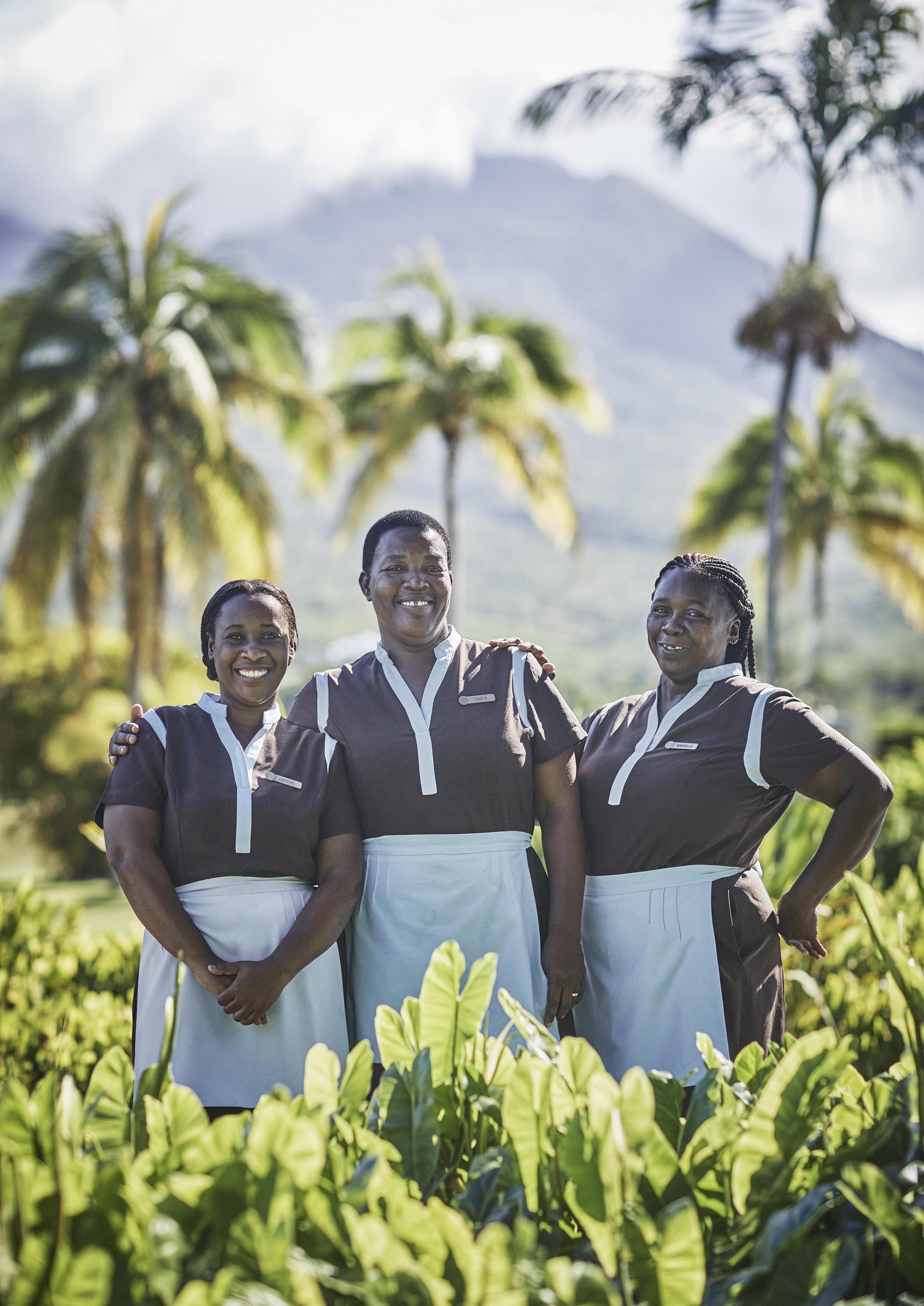 Three people in matching uniforms stand smiling in a lush garden with palm trees and a mountain in the background on a sunny day.