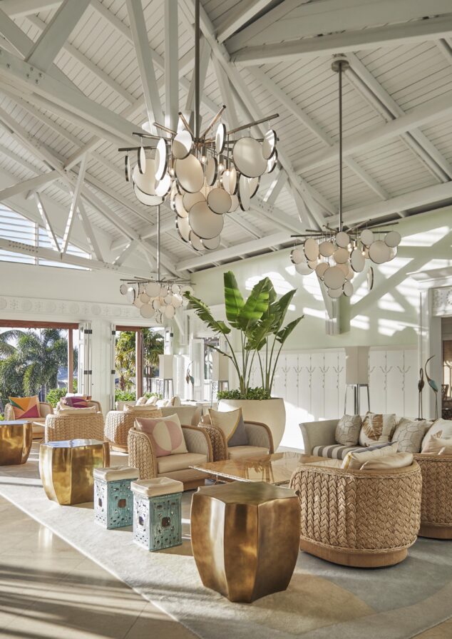 Spacious lobby with large windows. Features wicker chairs, gold accent tables, and large chandeliers. Green plants and outdoor view add to the bright and airy atmosphere.
