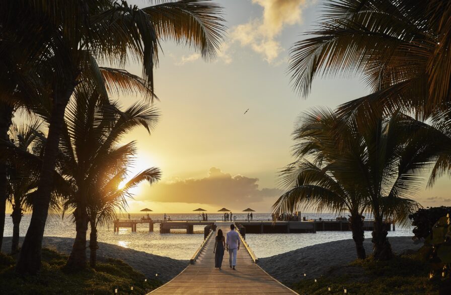 A couple walks on a wooden path towards a dock at sunset, surrounded by palm trees, with the ocean in the background.
