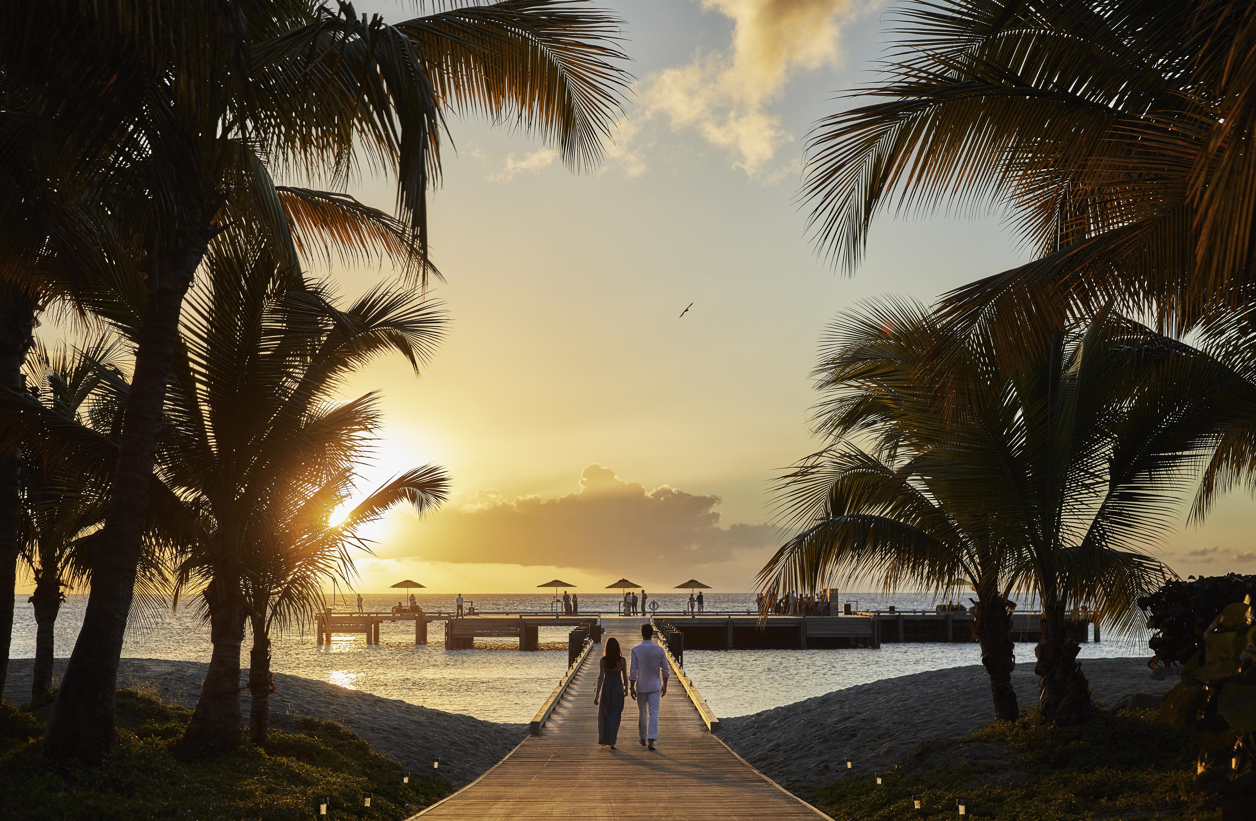 A couple walks on a wooden path towards a dock at sunset, surrounded by palm trees, with the ocean in the background.