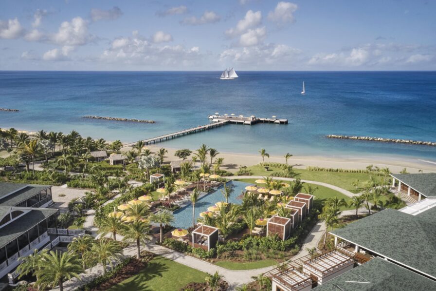 Aerial view of a coastal resort with a pool, yellow umbrellas, greenery, and a pier extending into the sea. Two sailboats are visible on the water under a partly cloudy sky.