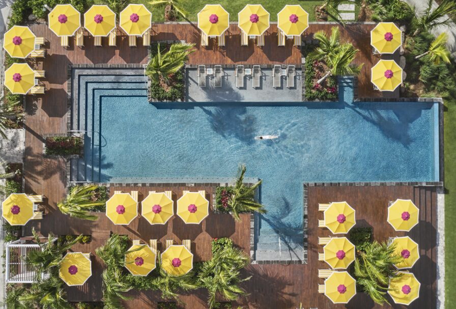 Aerial view of a rectangular pool with a swimmer, surrounded by wooden decking and yellow umbrellas. Palm trees are interspersed between seating areas.