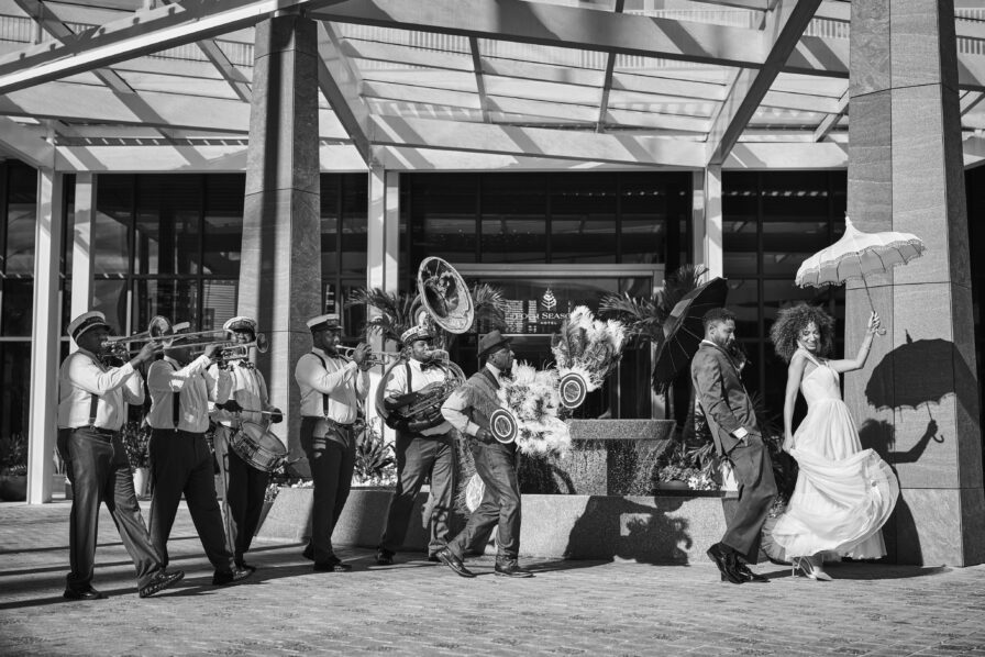 A couple dances outdoors in front of a marching band playing brass instruments. The woman holds an umbrella. The scene is lively and festive under a modern architectural structure.