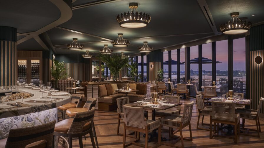 Elegant restaurant interior with wooden tables, woven chairs, and chandeliers. Large windows offer a view of the city skyline at dusk.