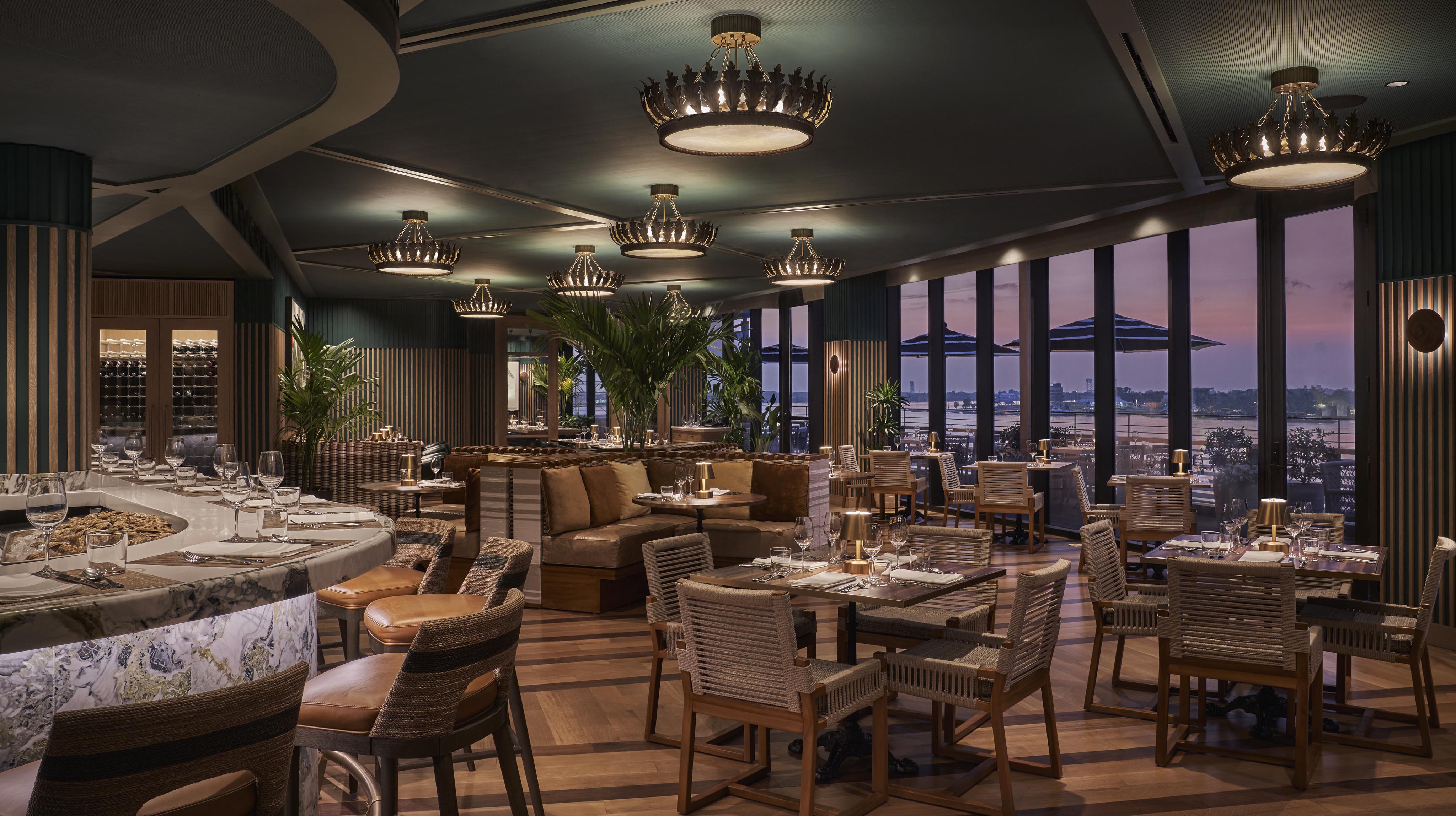Elegant restaurant interior with wooden tables, woven chairs, and chandeliers. Large windows offer a view of the city skyline at dusk.