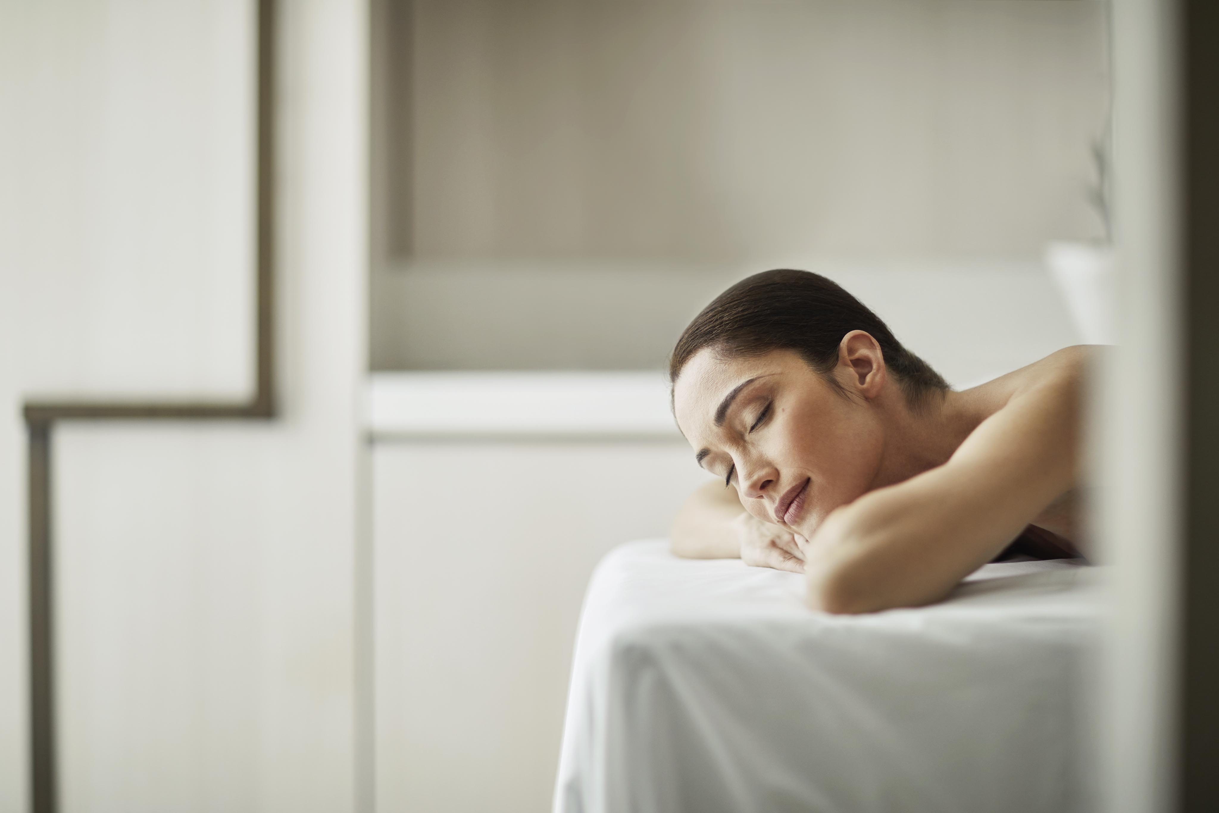 Person resting on a massage table with a serene expression, in a softly lit room with white decor.