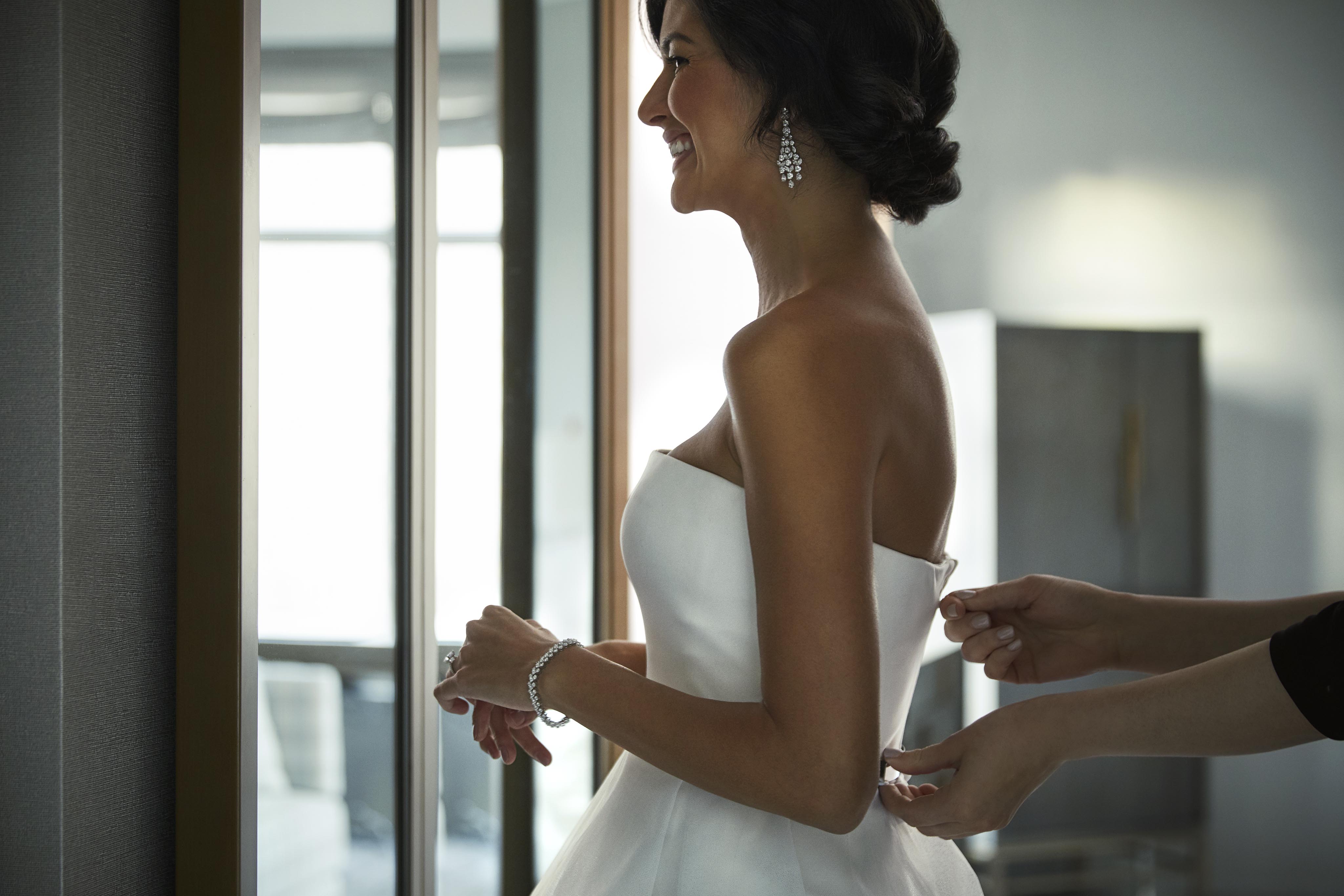 A woman in a white strapless dress is helped by another person, adjusting the back. She is smiling and wearing earrings and a bracelet.