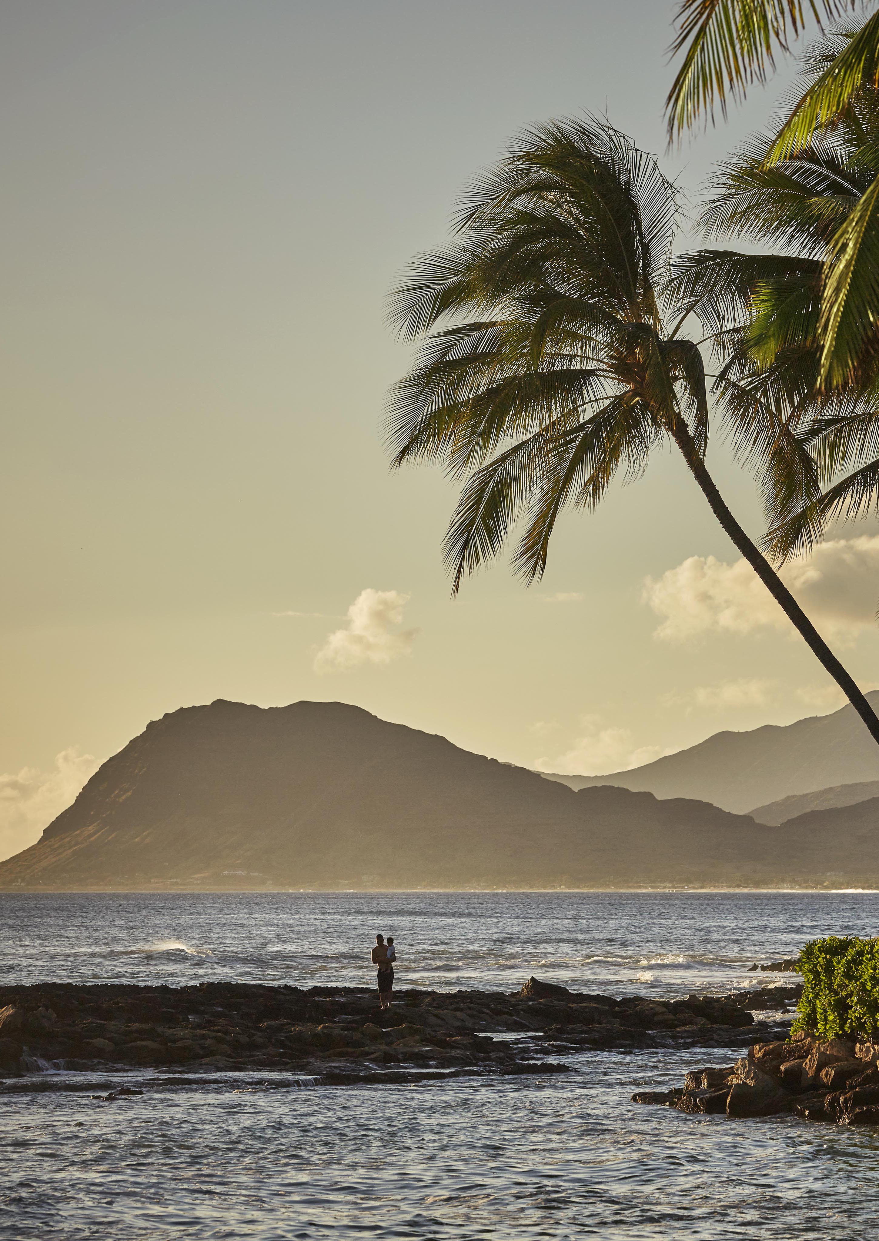 A person stands holding a child on rocky shore by the Lanikuhonua Oahu ocean at sunset, with lush tropical palm trees in the foreground and a mountain in the background.
