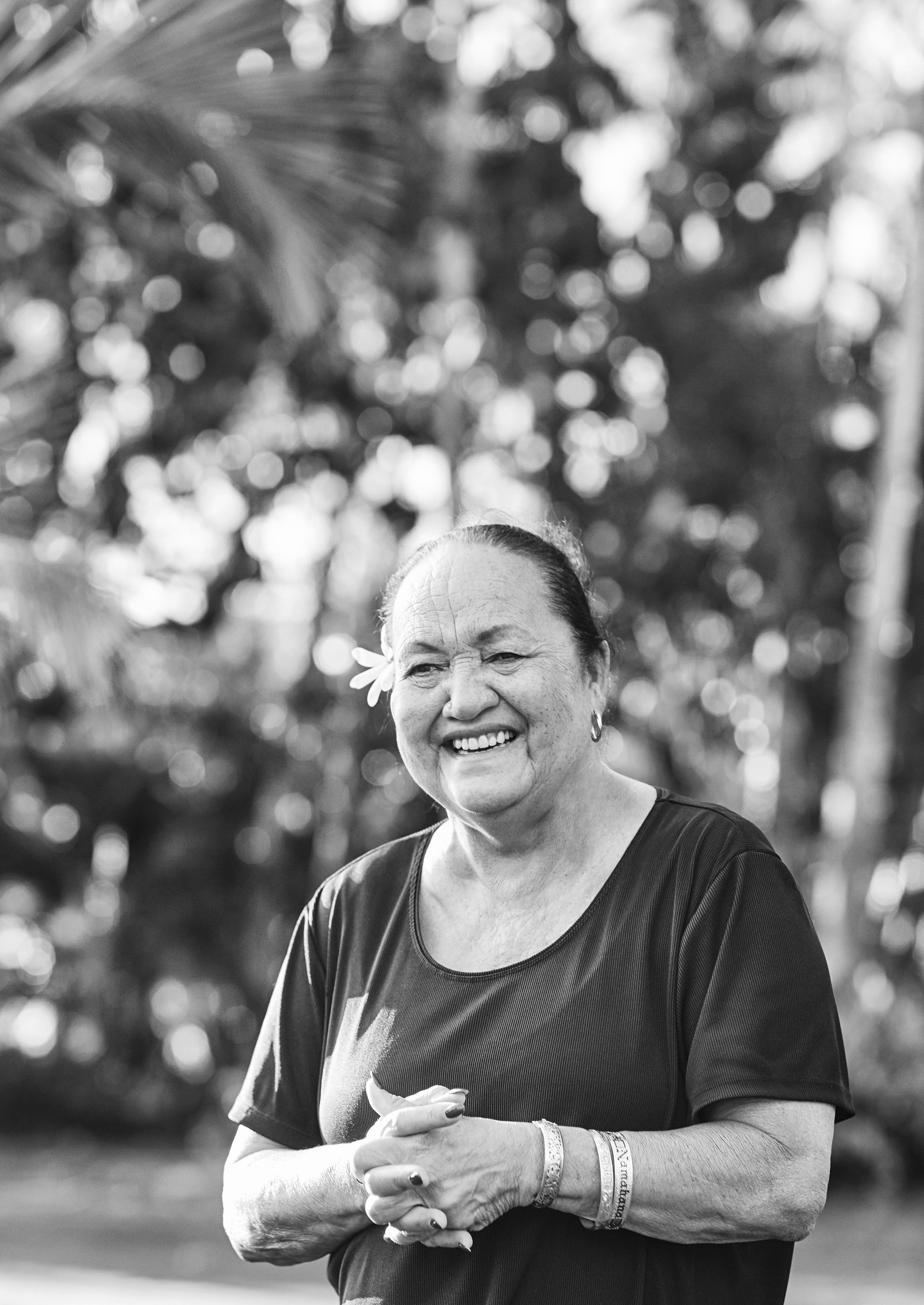An older woman photographed in black and white, wearing a flower behind her ear standing outdoors, smiling and holding her hands together. The background is blurred trees of Four Seasons Oahu property behind her.