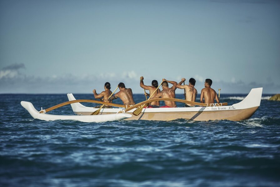 Six people paddle an outrigger canoe on the ocean under a clear sky.