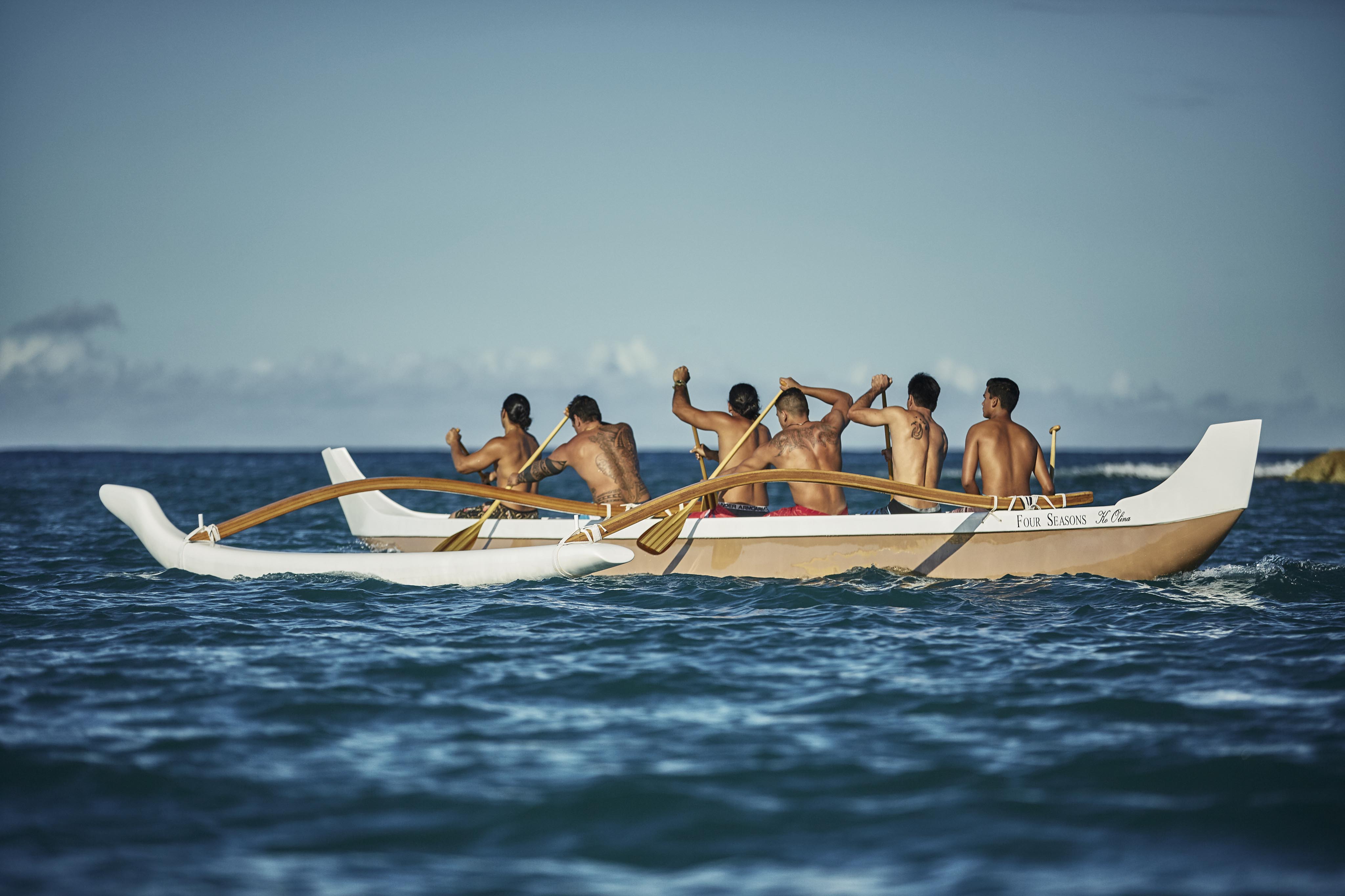 Six people paddle an outrigger canoe on the ocean under a clear sky.