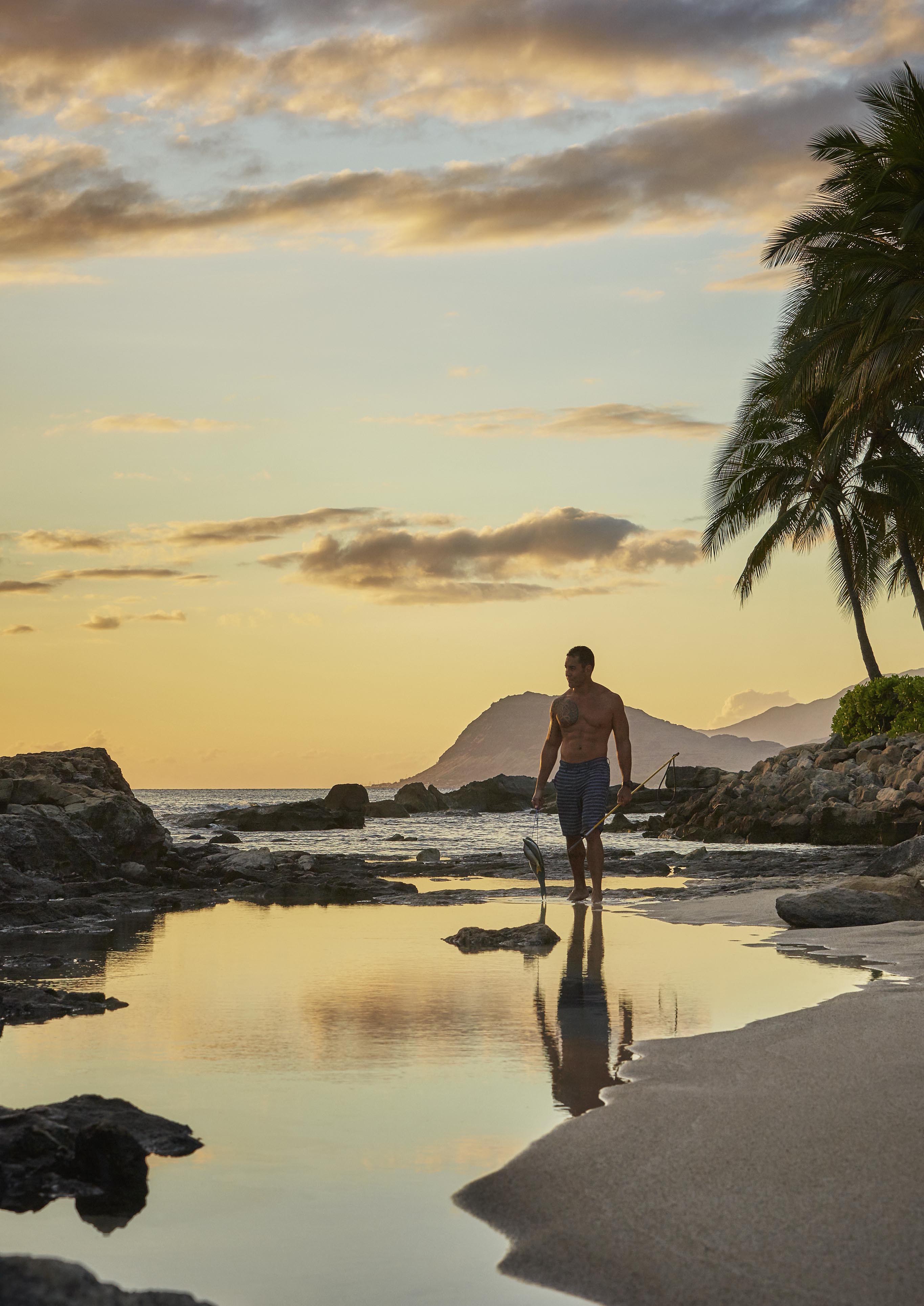 A local oahu spearfisherman stands shirtless on a beach at sunset, holding fresh caught fish and spear, with rocks, palm trees, and hawaiian mountains in the background.