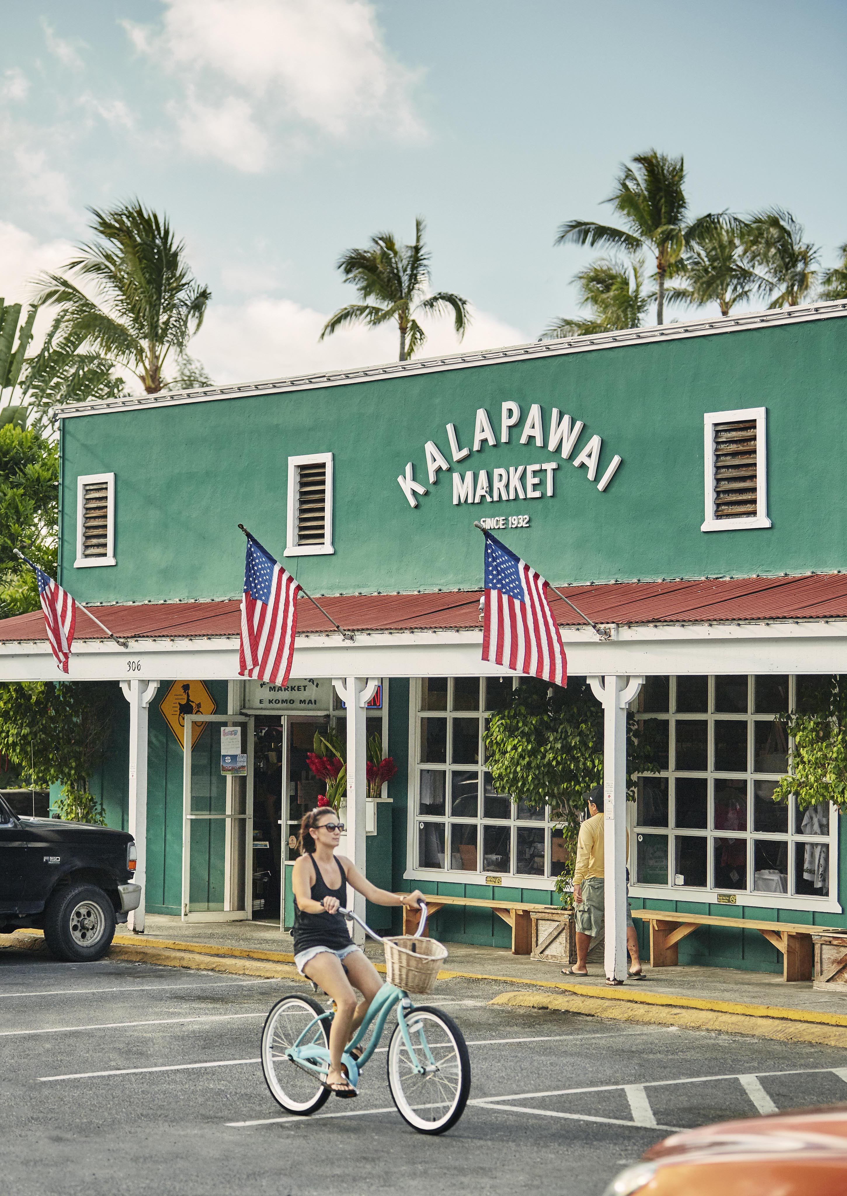 A person rides a bicycle past the vibrant green Kalapawai Market building on the North Shore of Hawaii, the exterior decorated with American flags and surrounded by palm trees.