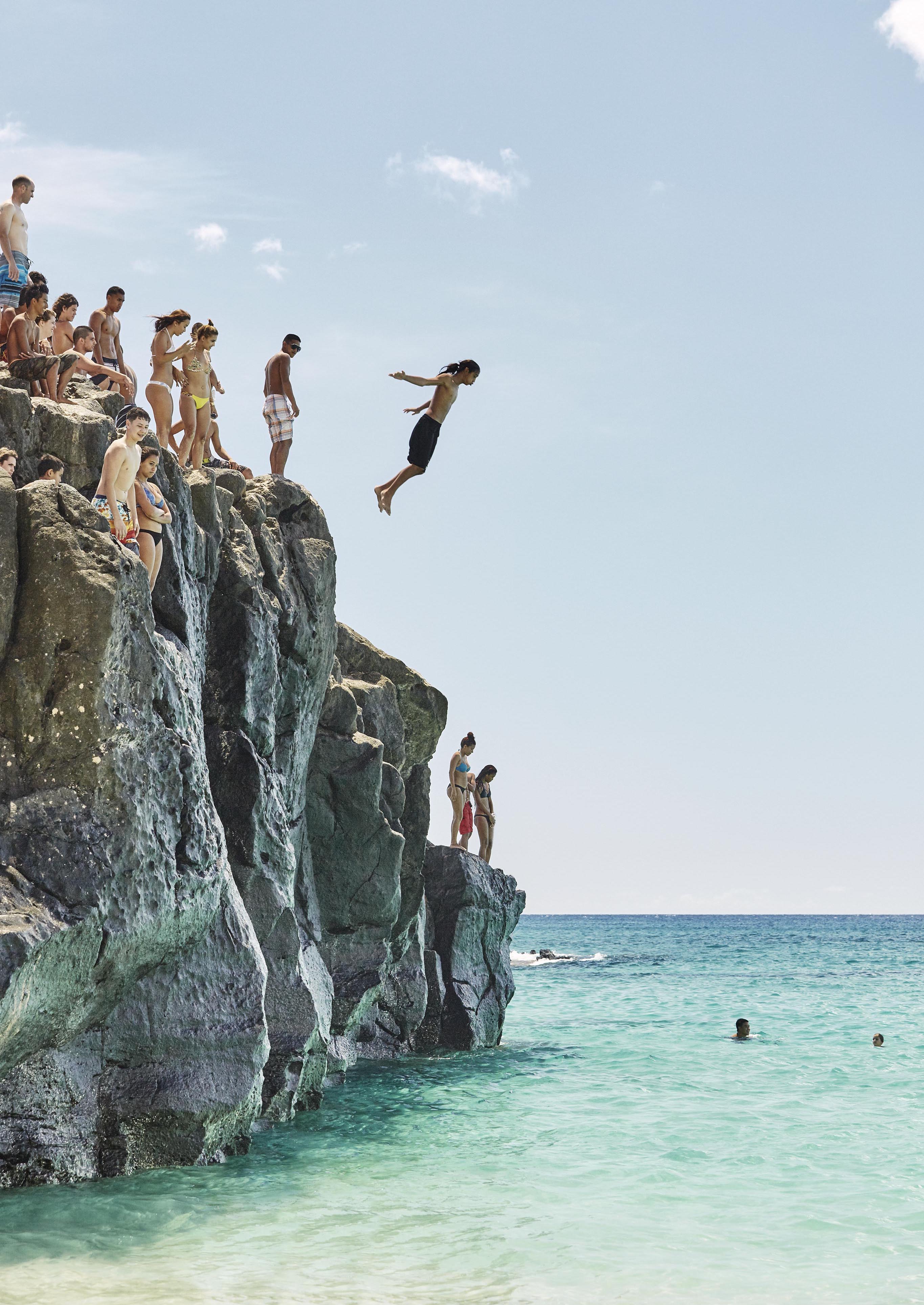 A group of young people stand on rocky cliffs by the north shore ocean while one person jumps off into the Oahu, Hawaii water below.