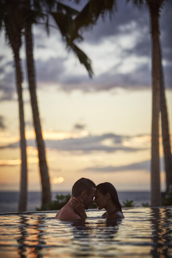 A couple embraces in a pool at sunset, with palm trees and the ocean in the background.