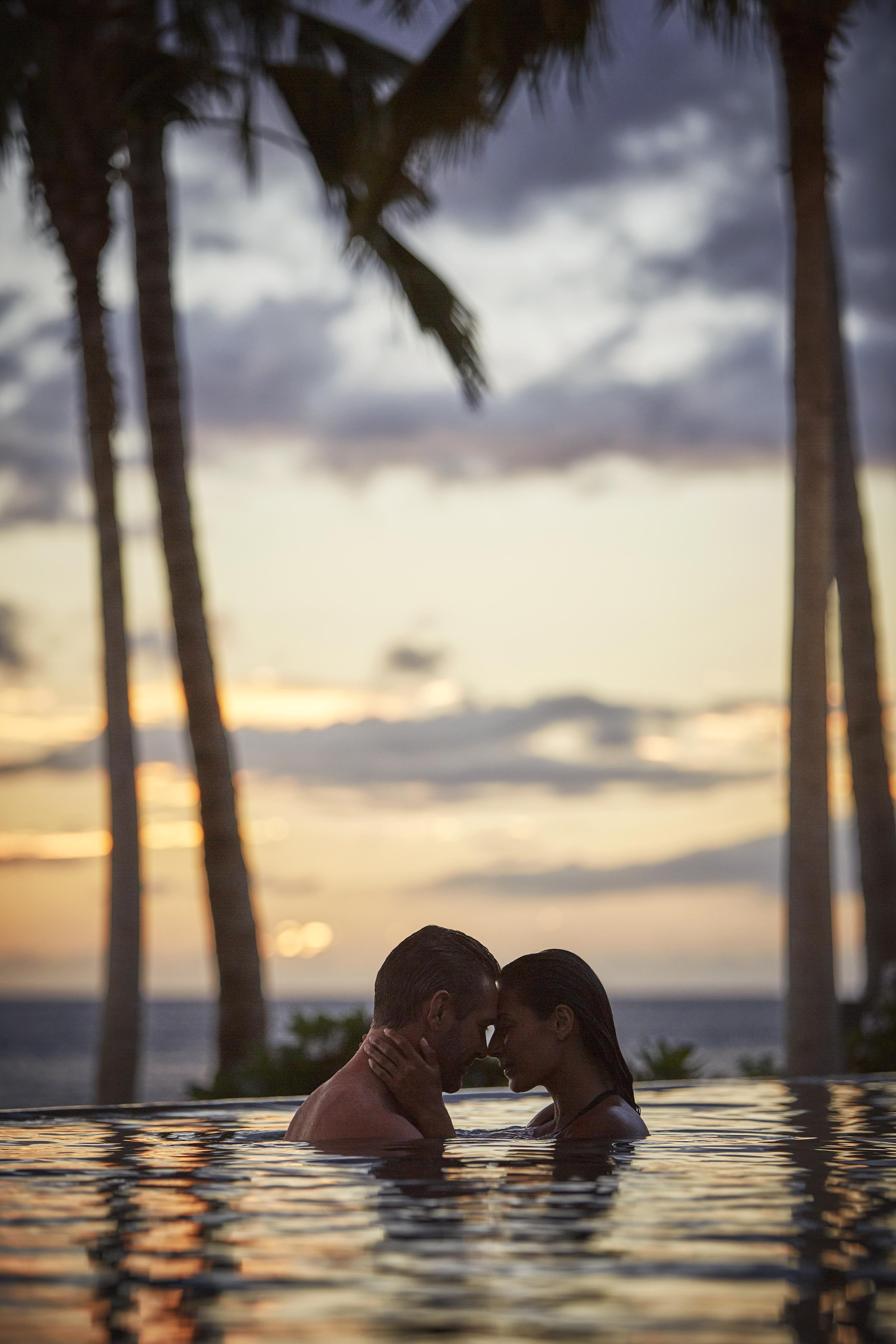 A couple embraces in a pool at sunset, with palm trees and the ocean in the background.