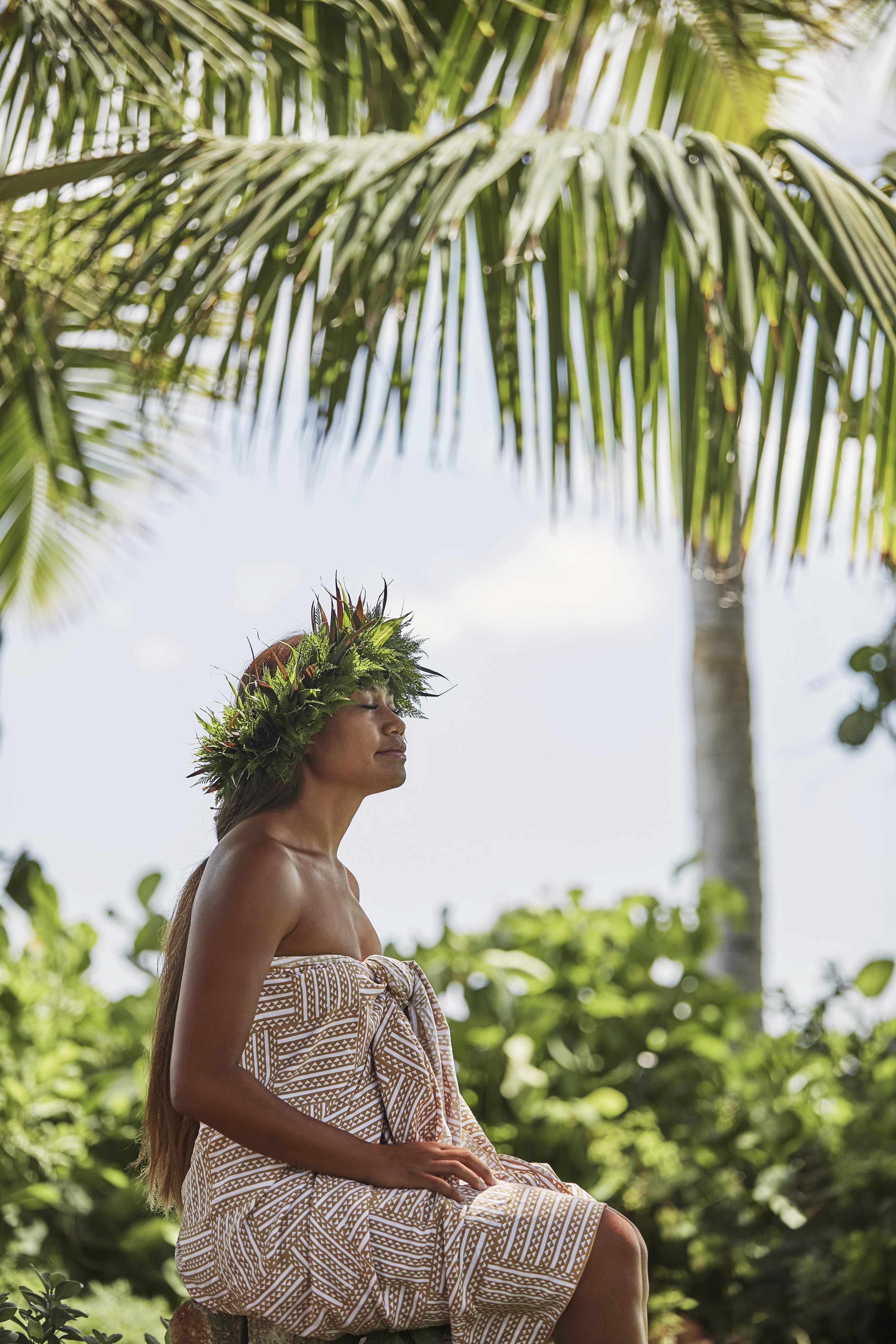 A woman wearing a leaf crown and patterned outfit sits outdoors under a palm tree on a sunny day.
