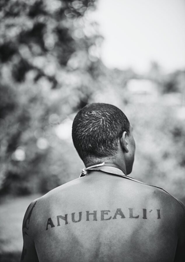 A person with short hair is seen from behind, showing a tattoo on their upper back that reads "ANUHEALI'I." The background is blurred greenery.