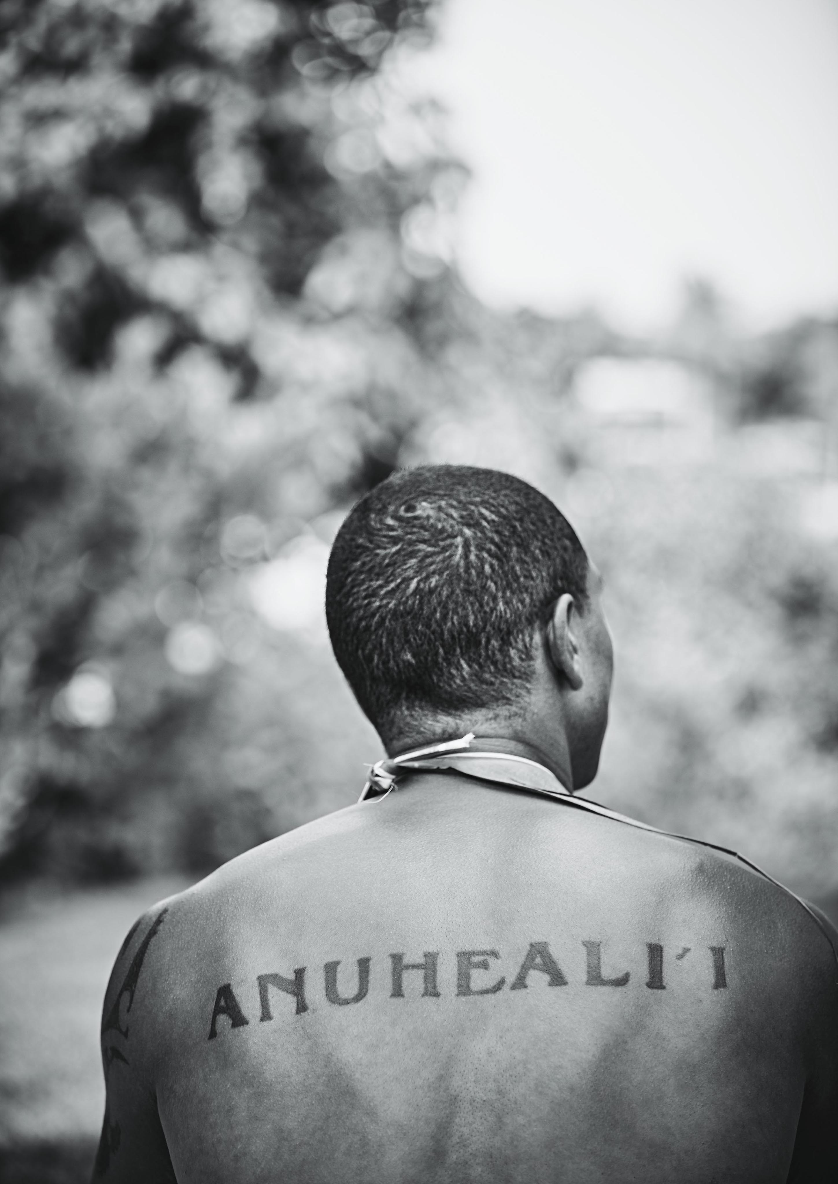 A person with short hair is seen from behind, showing a tattoo on their upper back that reads "ANUHEALI'I." The background is blurred greenery.