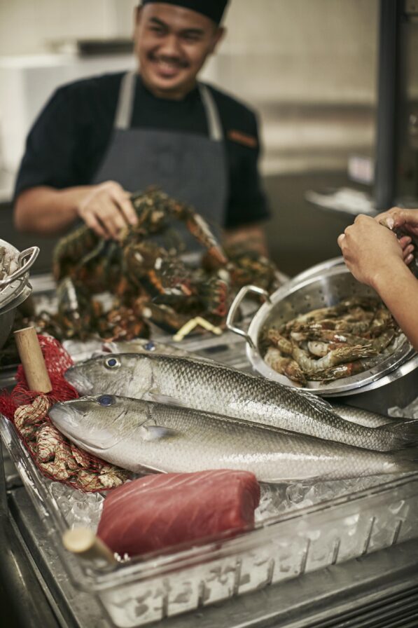 A chef handling lobsters and prawns near a tray with two large fish and a piece of raw tuna on ice. A person is holding a pot of prawns.