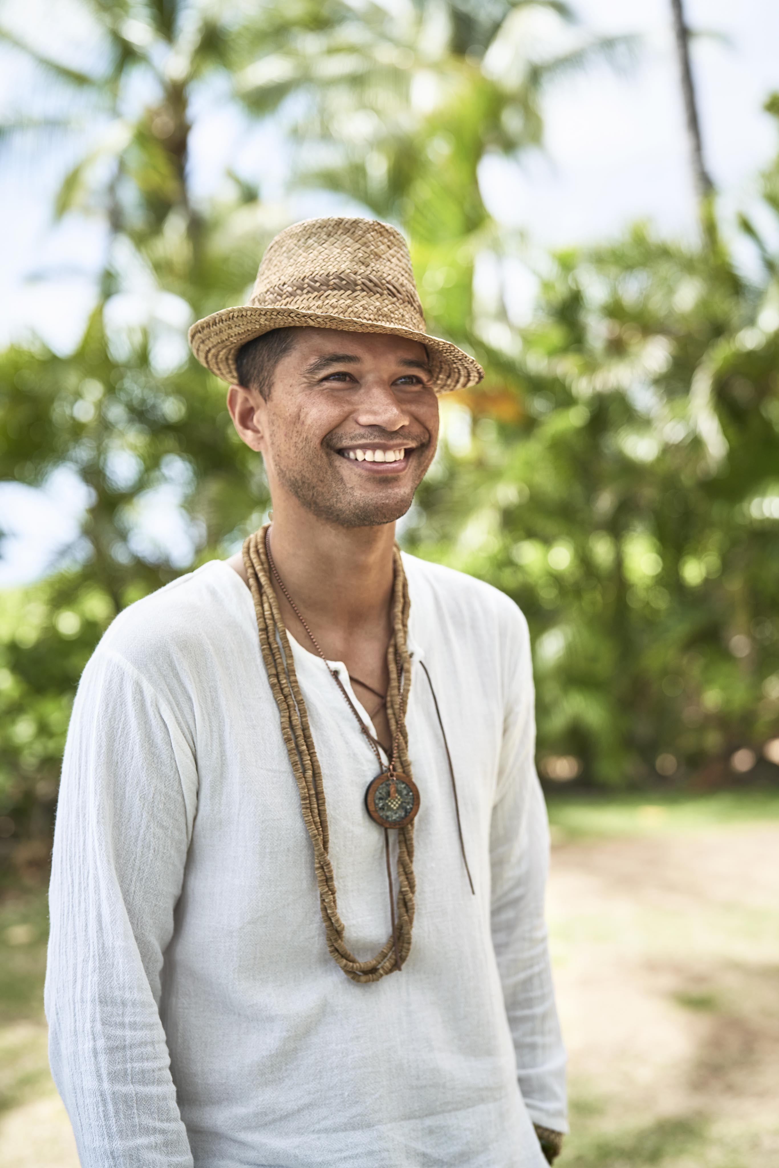Lifestyle capture of man in a white shirt and straw hat stands outdoors, smiling. They are wearing a beaded necklace. Palm trees and greenery are in the background of Four Seasons Oahu.