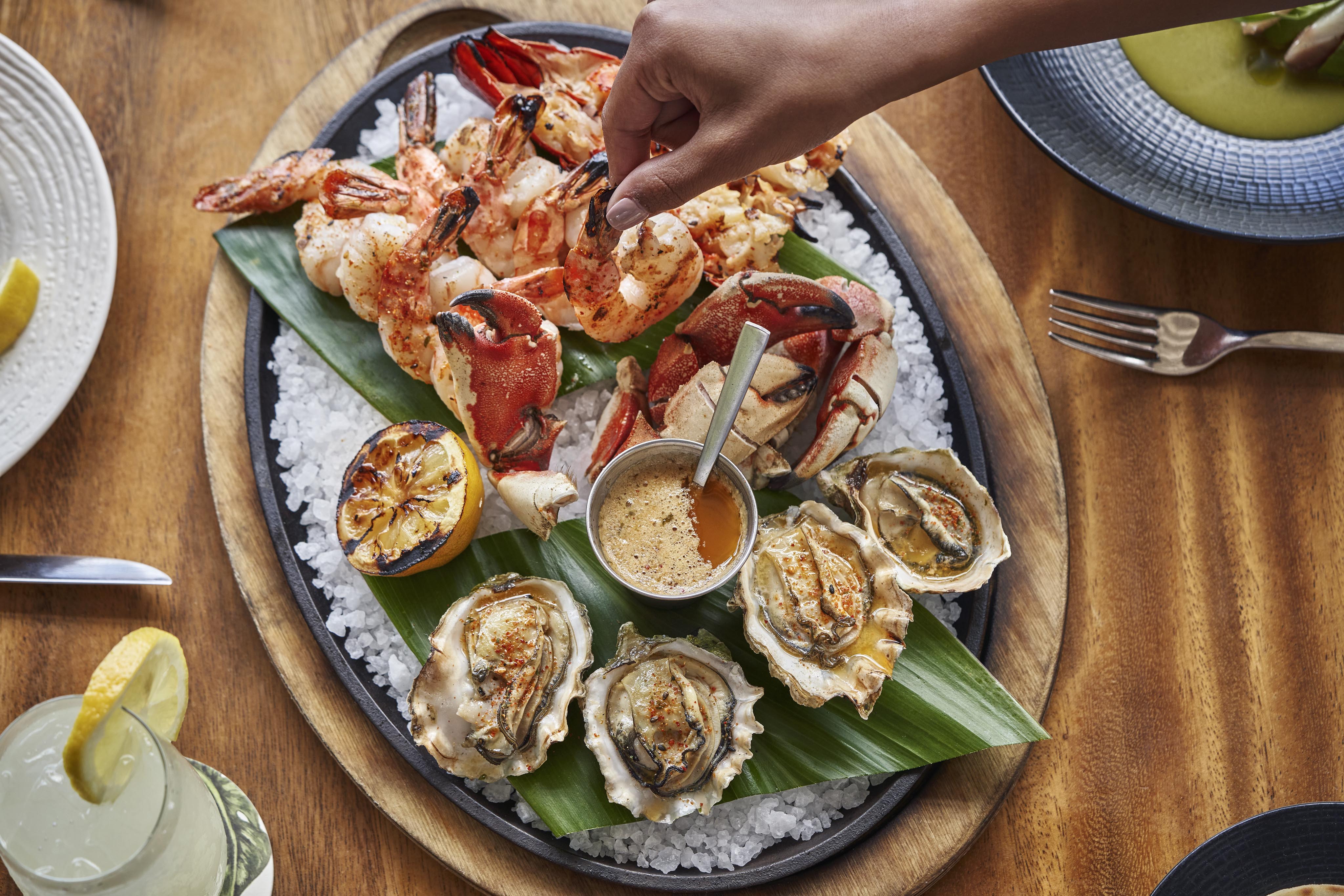 A seafood platter featuring shrimp, crab, oysters, grilled lemon, and dipping sauce on a bed of ice. A hand reaches for a shrimp. Plates and a drink surround the wooden serving tray.