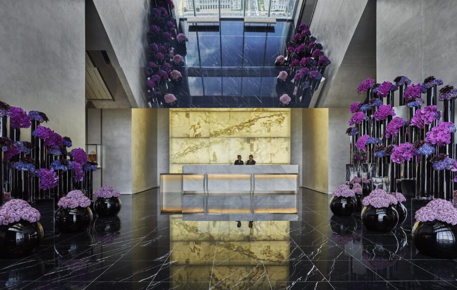 Luxury resort lobby of Four Seasons Philadelphia with two people at a reception desk. Marble flooring reflects purple floral arrangements and a gold-accented wall. Large ceiling mirrors enhance the spacious design.