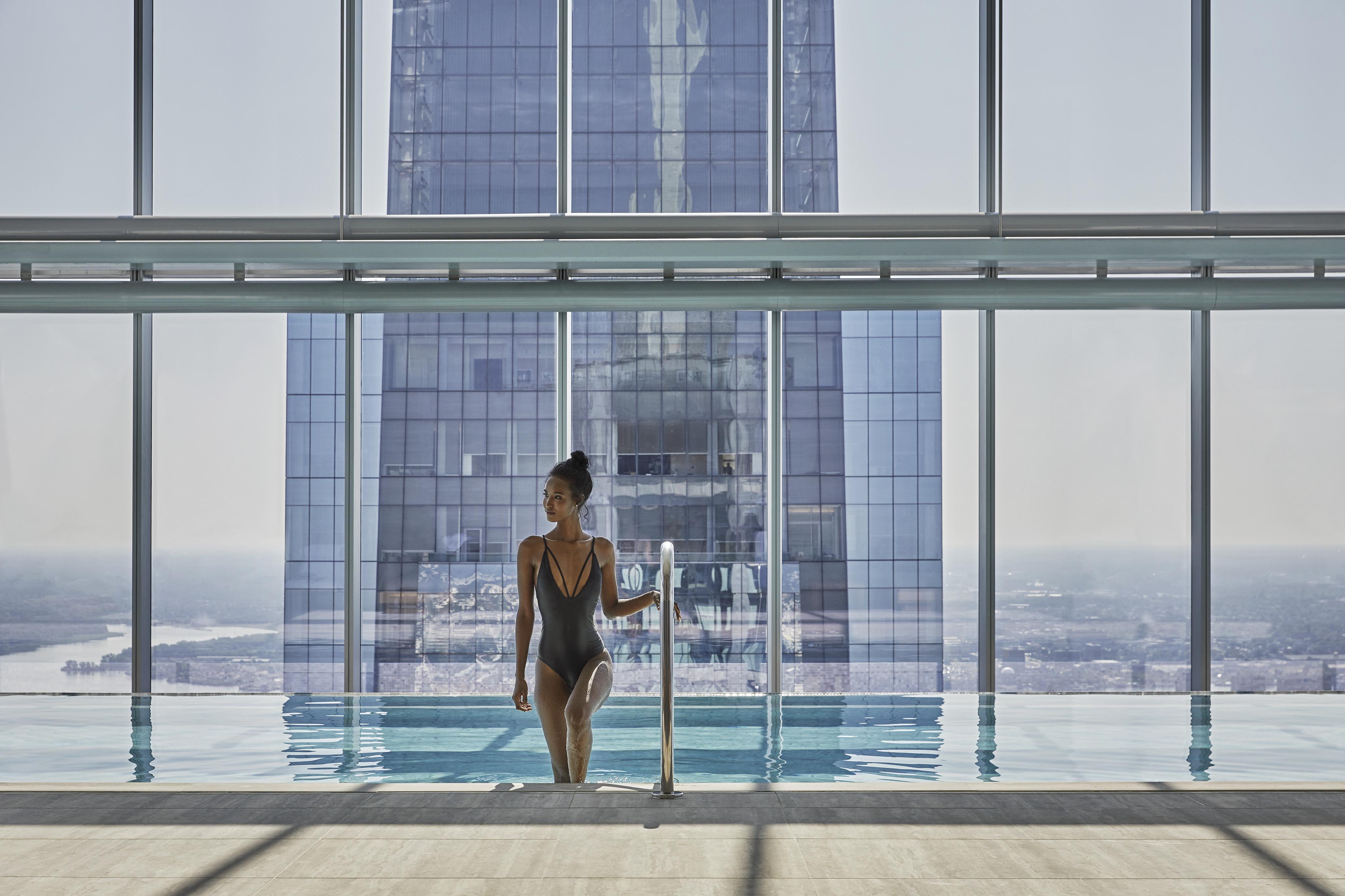 Woman steps into an indoor pool with large windows showing a cityscape and a tall glass building in the background.