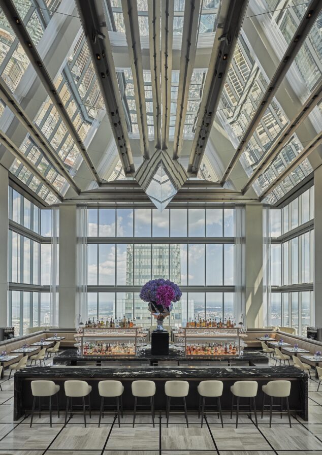 Spacious rooftop bar of luxury hotel with high glass windows and a central black marble counter surrounded by beige stools. A large floral arrangement sits atop the bar against a backdrop of cityscape views.