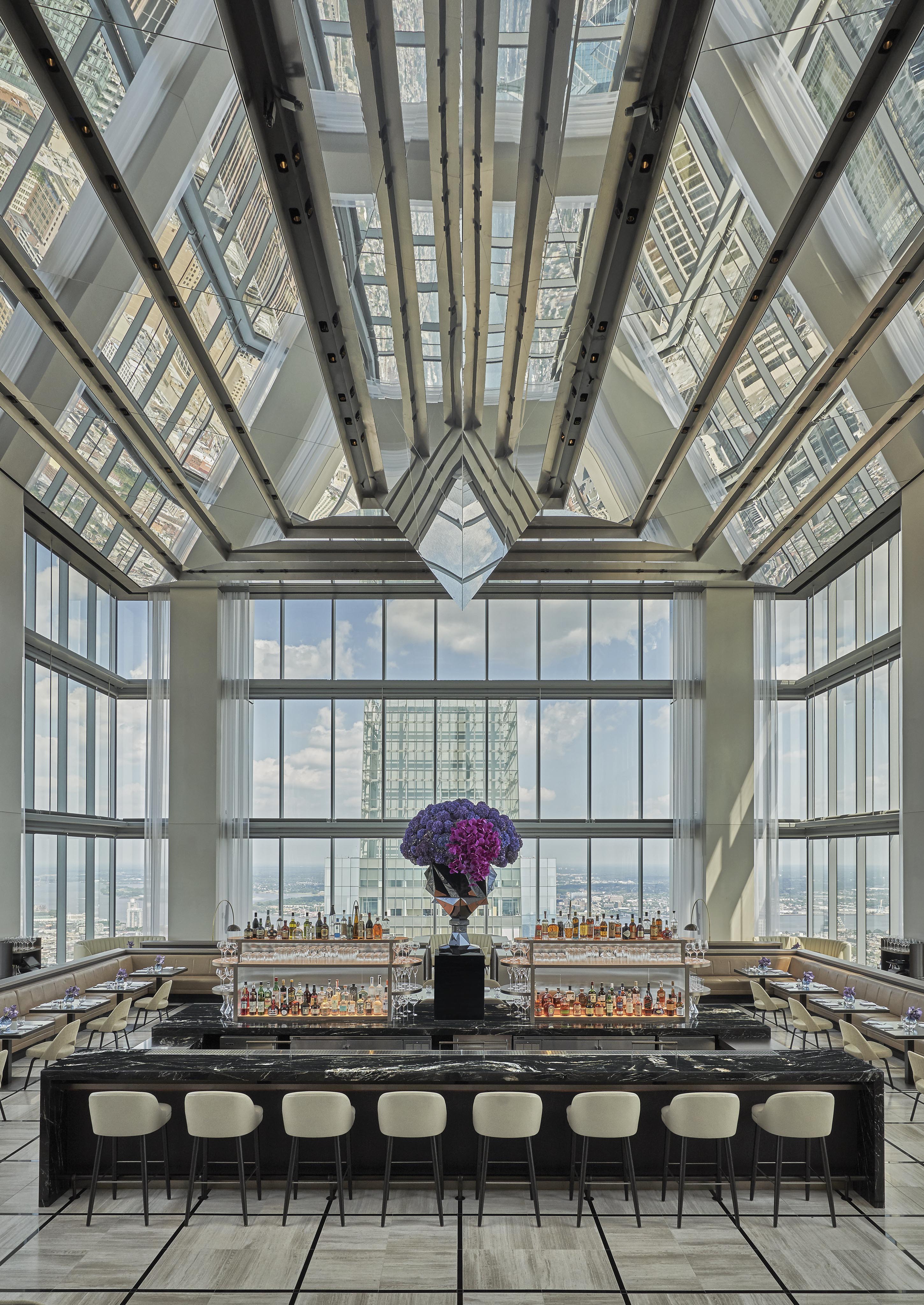 Spacious rooftop bar of luxury hotel with high glass windows and a central black marble counter surrounded by beige stools. A large floral arrangement sits atop the bar against a backdrop of cityscape views.