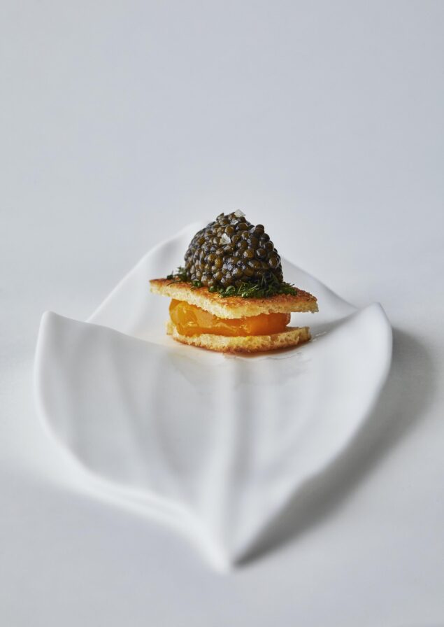 An elegant and detailed capture of elevated gourmet appetizer of caviar on a wafer, garnished with herbs, and placed on a white, leaf-shaped plate.