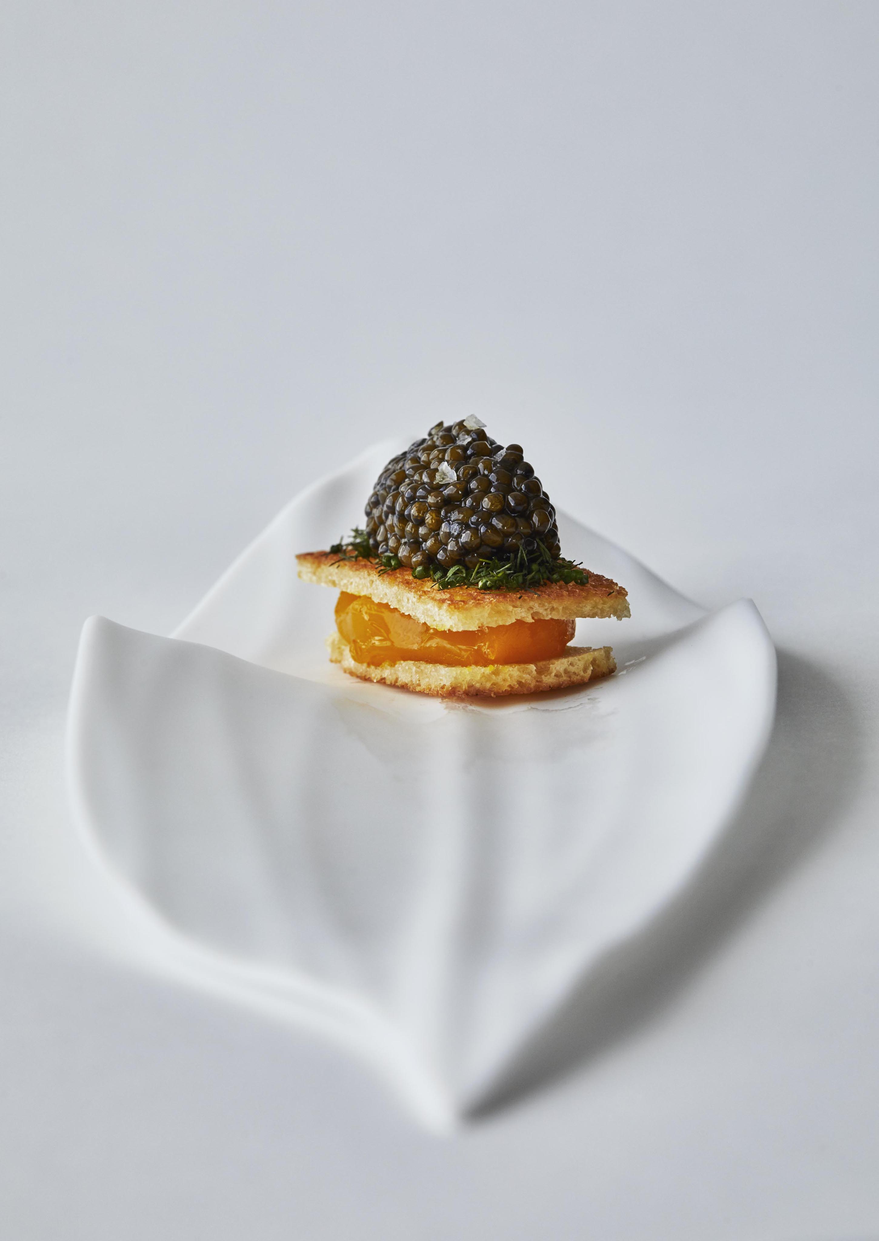 An elegant and detailed capture of elevated gourmet appetizer of caviar on a wafer, garnished with herbs, and placed on a white, leaf-shaped plate.