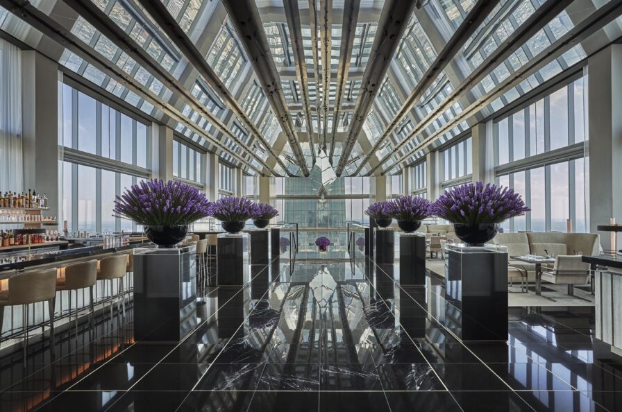 Modern rooftop bar of Four Seasons Philadelphia hotel with floor-to-ceiling windows, glass ceiling, black reflective floor, and purple floral arrangements. Bar on the left, seating on the right.