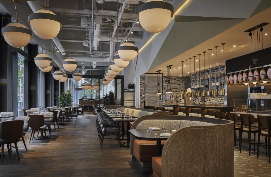 An architecture view of modern Four Seasons Philadelphia hotel restaurant interior with round pendant lights, wooden floors, and cushioned seating. Tables are set with plates and utensils. Large windows and plants are visible in the background.