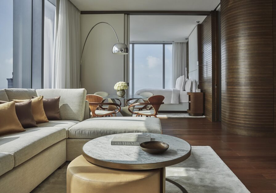 A modern Four Seasons Philadelphia luxury hotel interior with a beige sectional sofa, round marble coffee table, and curved floor lamp. A small dining area with wooden chairs is in the background, near a large window.