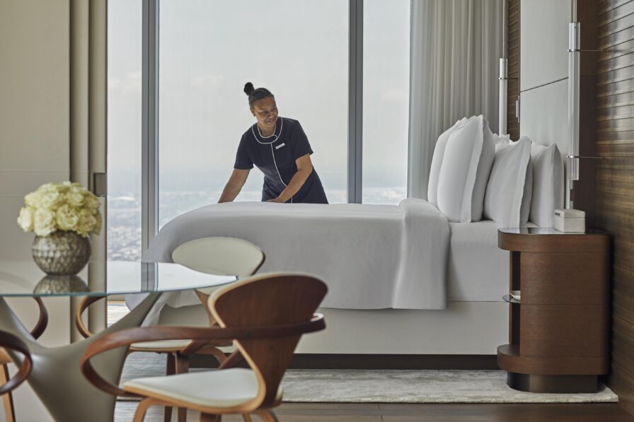 Modern hotel room architecture of Four Seasons Philadelphia featuring a neatly made bed near a large window, with a view of the city. A glass table with wooden chairs and a vase of flowers is in the foreground.