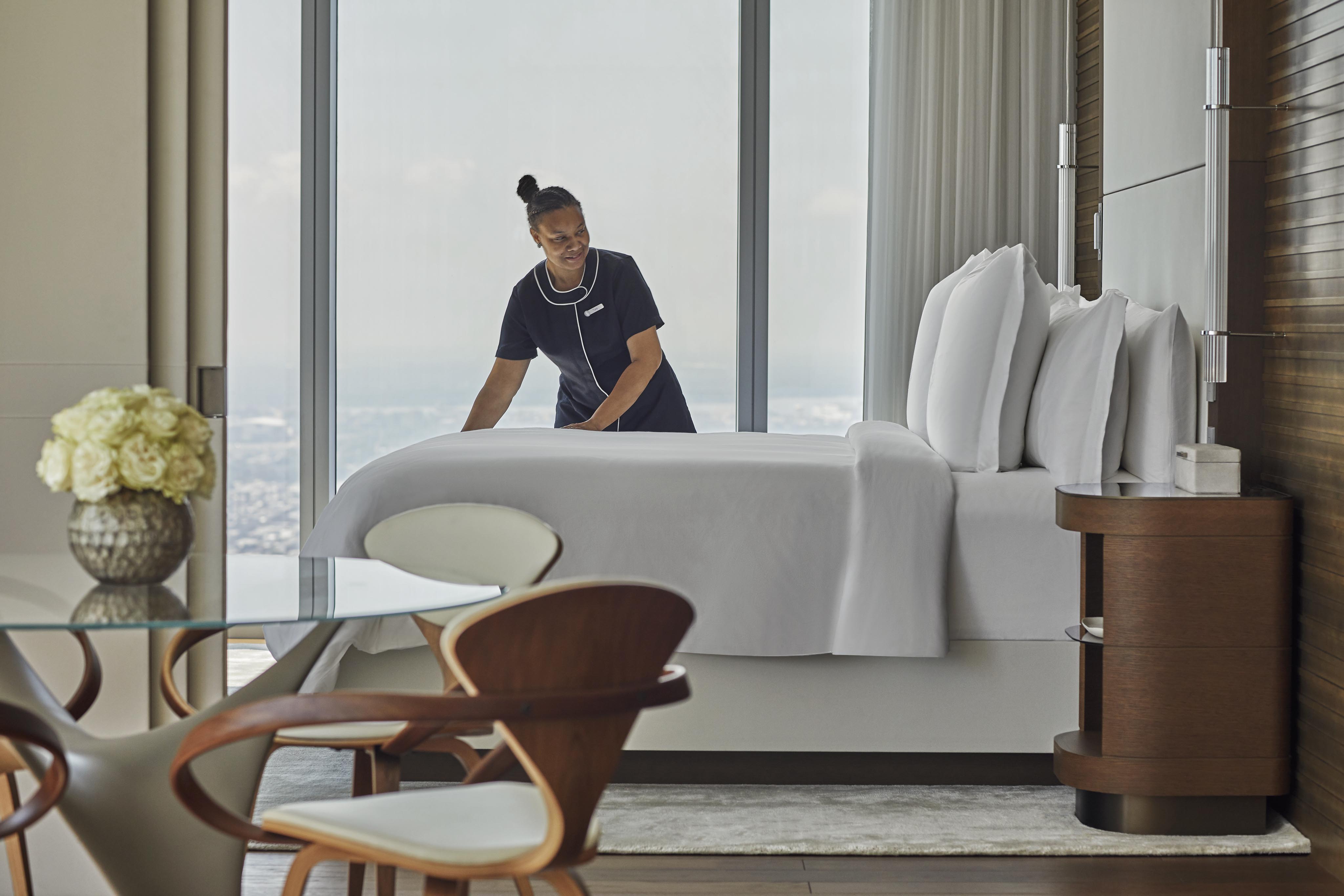 Modern hotel room architecture of Four Seasons Philadelphia featuring a neatly made bed near a large window, with a view of the city. A glass table with wooden chairs and a vase of flowers is in the foreground.