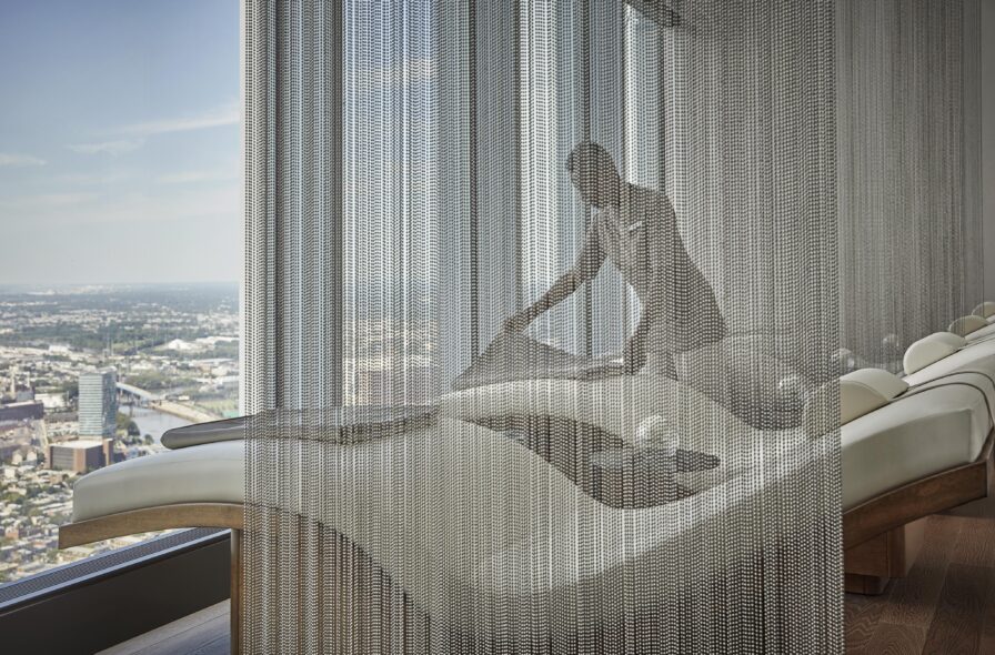 A person adjusts seating in a modern, high-rise luxury resort and spa lounge featuring sleek, curved chairs and a window view of an urban landscape through chain-like partitions.