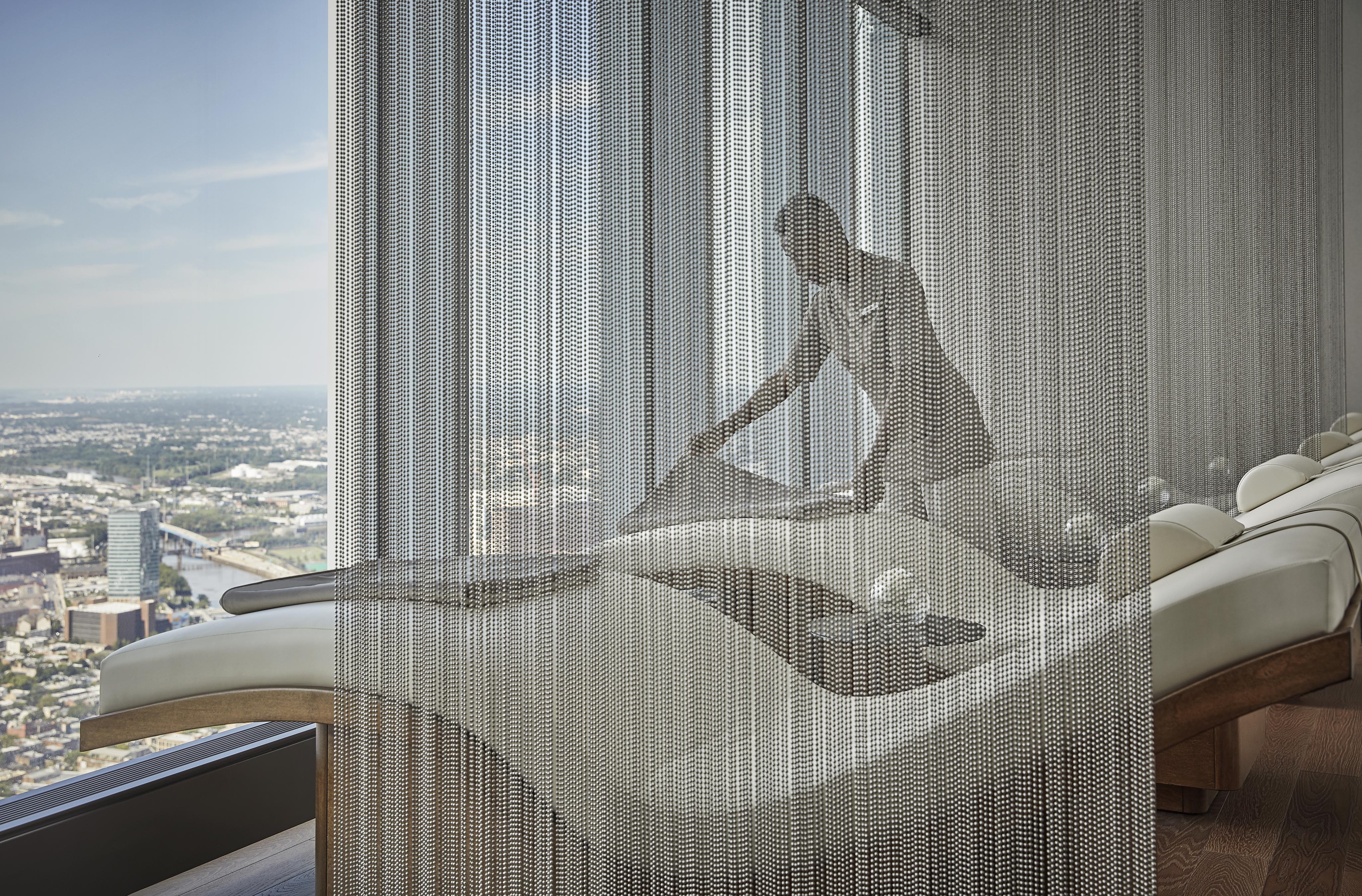 A person adjusts seating in a modern, high-rise luxury resort and spa lounge featuring sleek, curved chairs and a window view of an urban landscape through chain-like partitions.