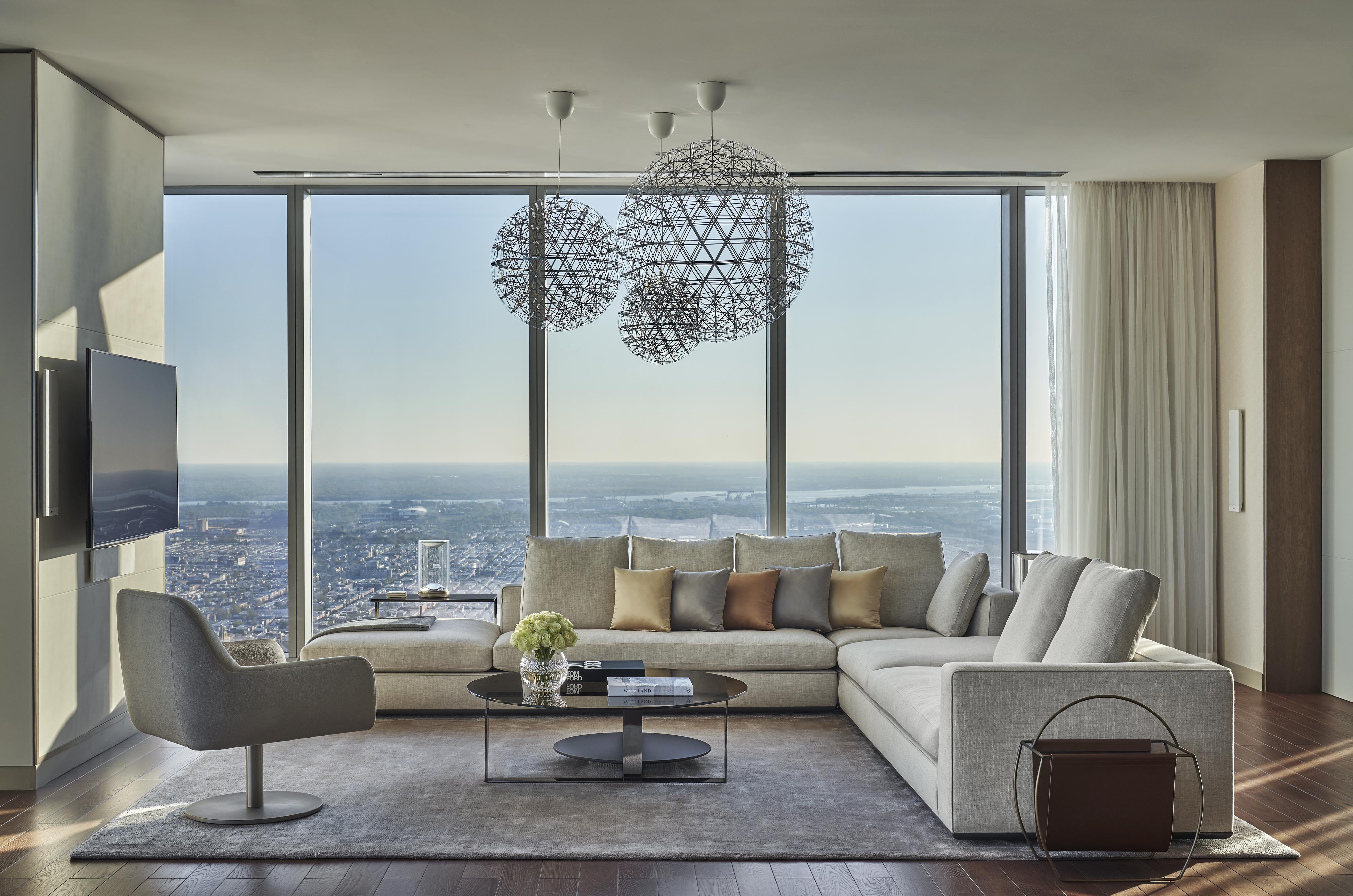 Pristine Four Seasons Philadelphia hotel suite interior with a large gray sectional sofa, round glass coffee table, and swivel chair. Floor-to-ceiling windows offer a city view. Geometric pendant lights hang from the ceiling.