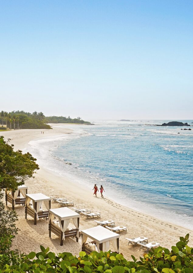 A couple walking the Four Seasons Punta Mita resort beach lined with exclusive luxury resort cabanas surrounded by lush greenery.