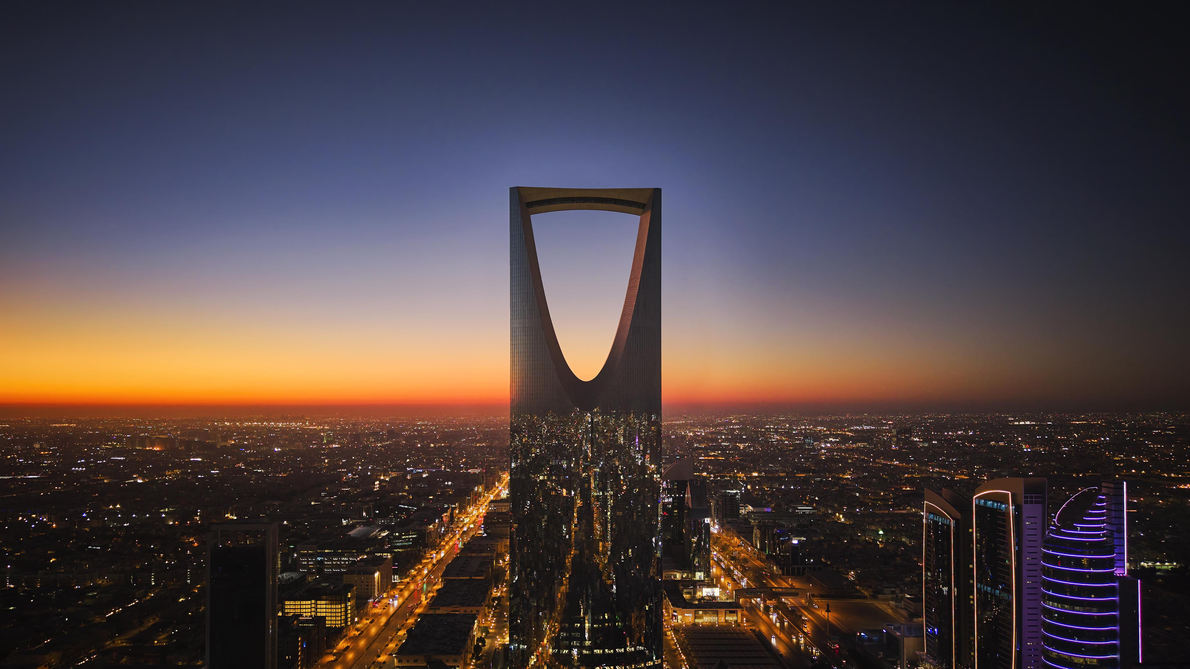 A tall, modern skyscraper—possibly a luxury hotel and resort—features a distinctive oval cutout at the top, standing in the center of a Middle East cityscape at sunset, lights illuminating the buildings below.