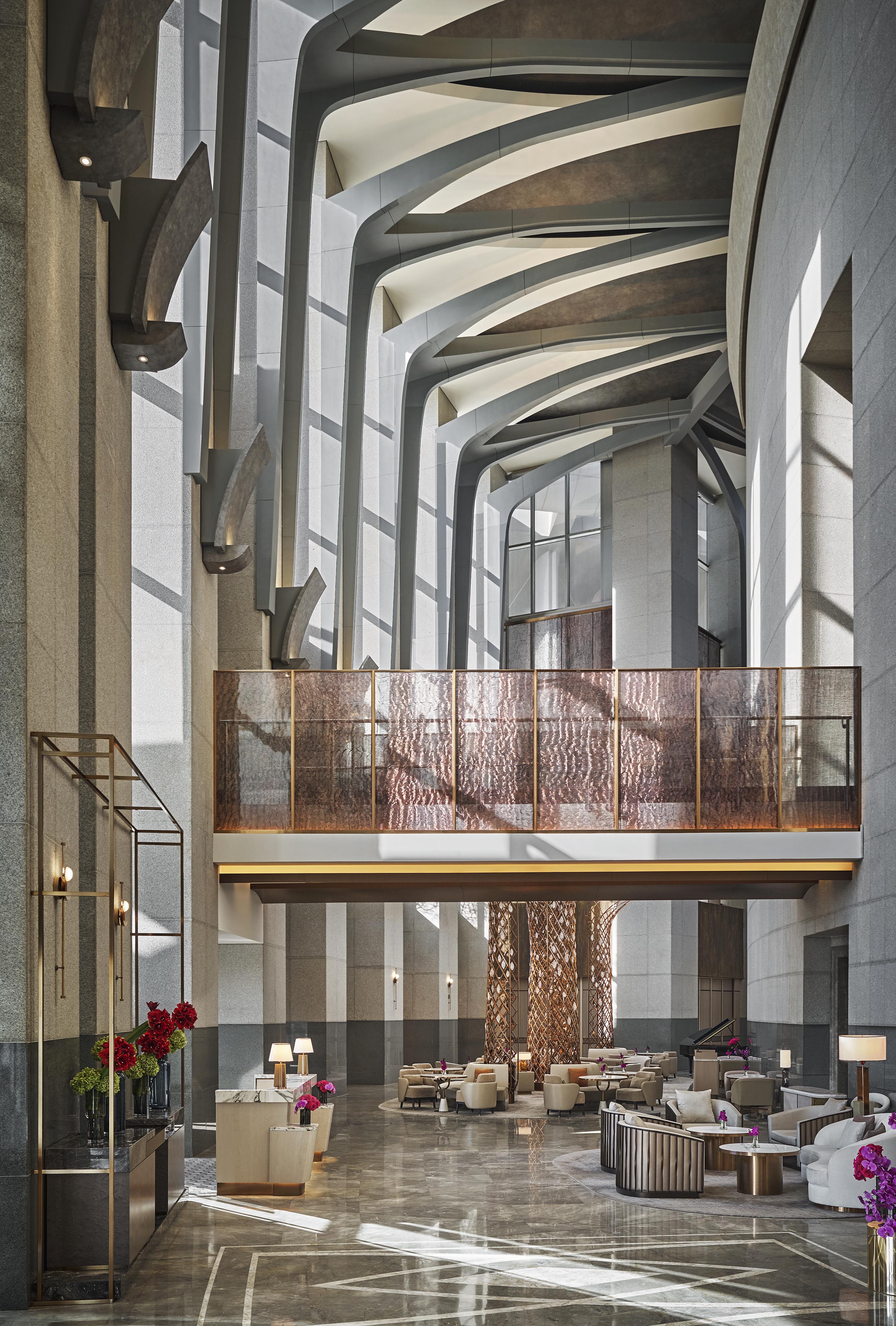 A spacious, modern luxury hotel and resort lobby with high ceilings, large windows, sculptural beams, elegant seating areas, and floral arrangements—perfect for Middle East photography.