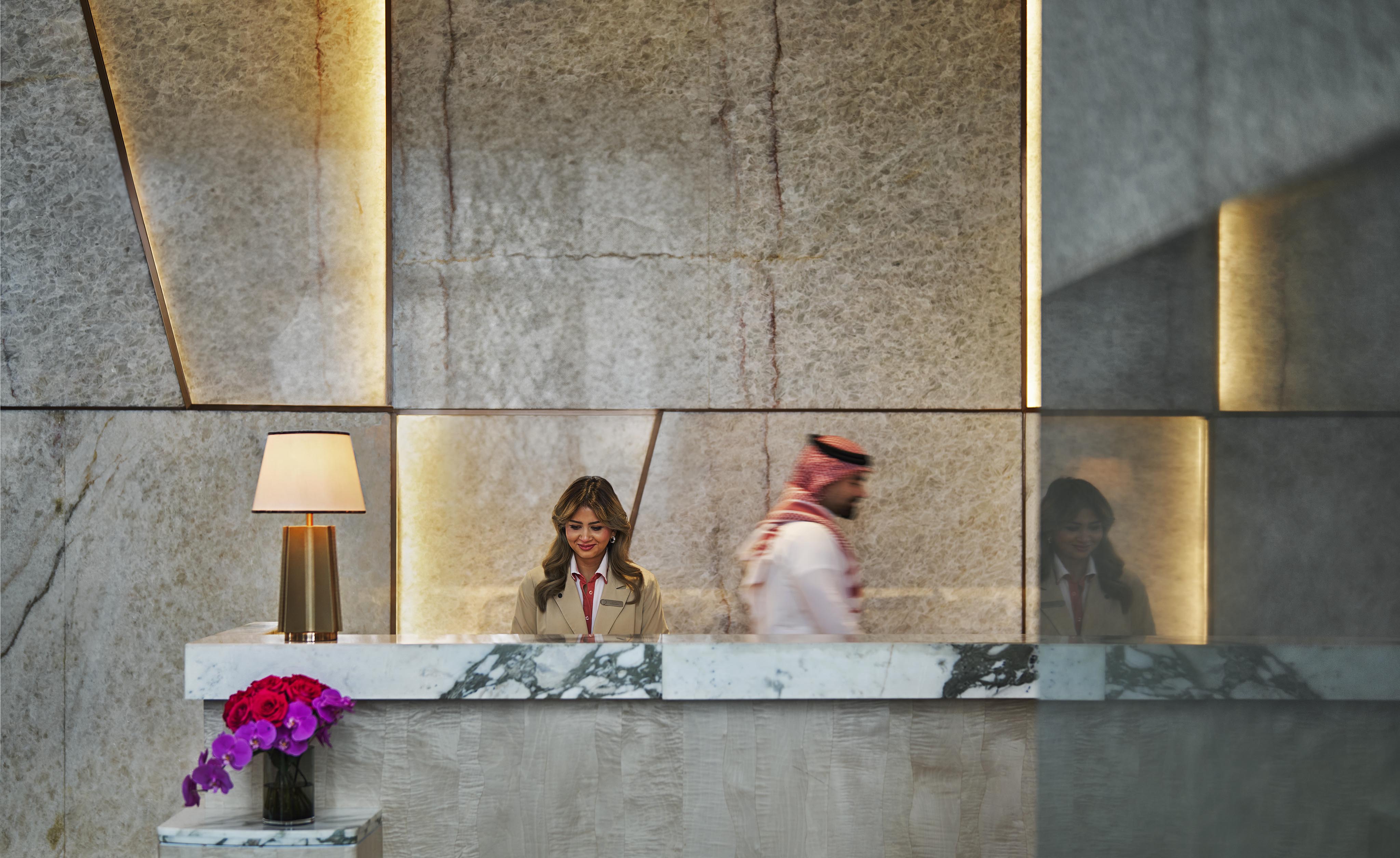 A woman stands behind a marble reception desk with a lamp and flowers nearby, while a man in traditional Middle Eastern attire walks past—capturing the elegance of luxury hotel and resort photography in the Middle East.