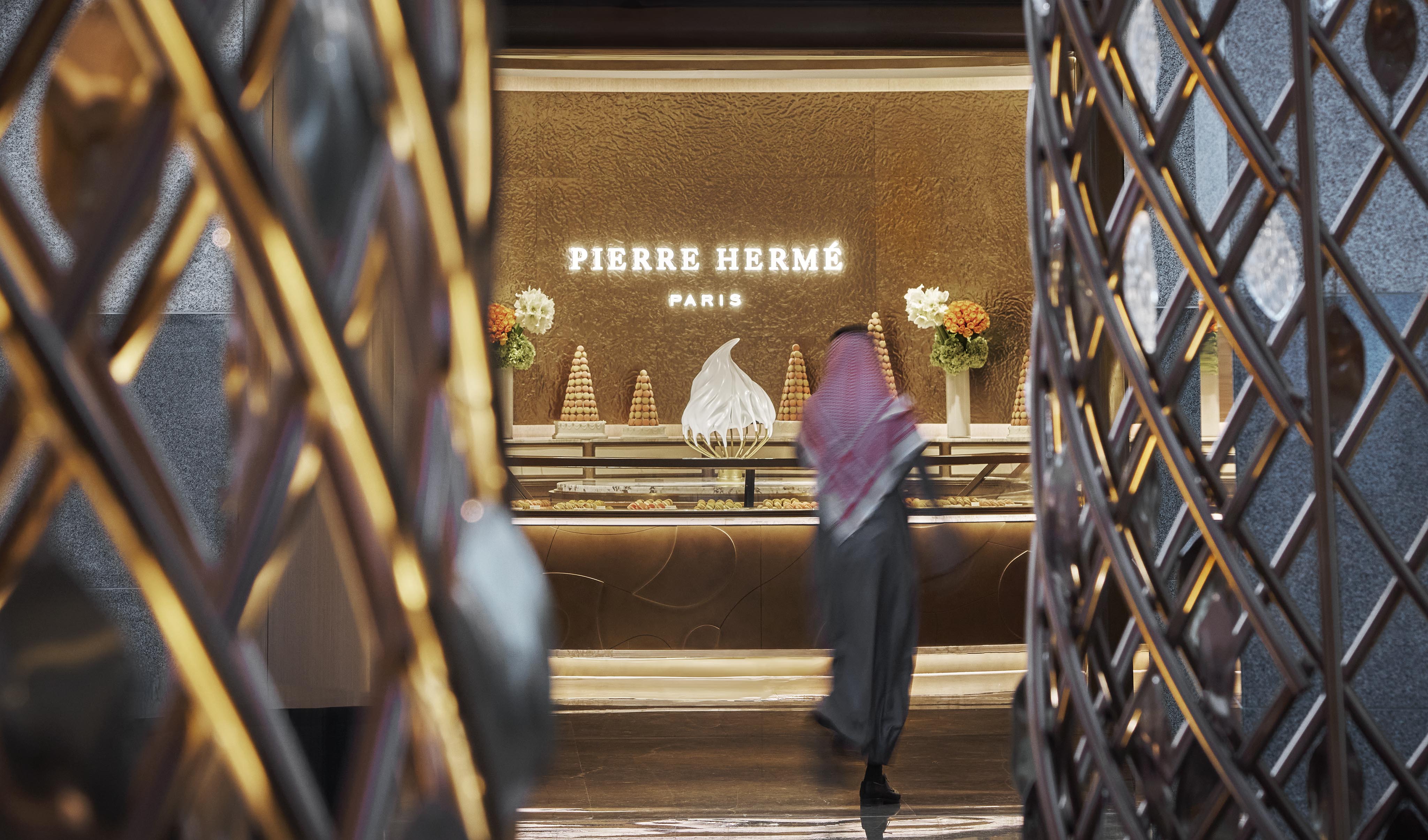 A person in traditional attire stands in front of the Pierre Hermé Paris counter at a luxury hotel and resort in the Middle East, with exquisite desserts and floral arrangements displayed on the counter.