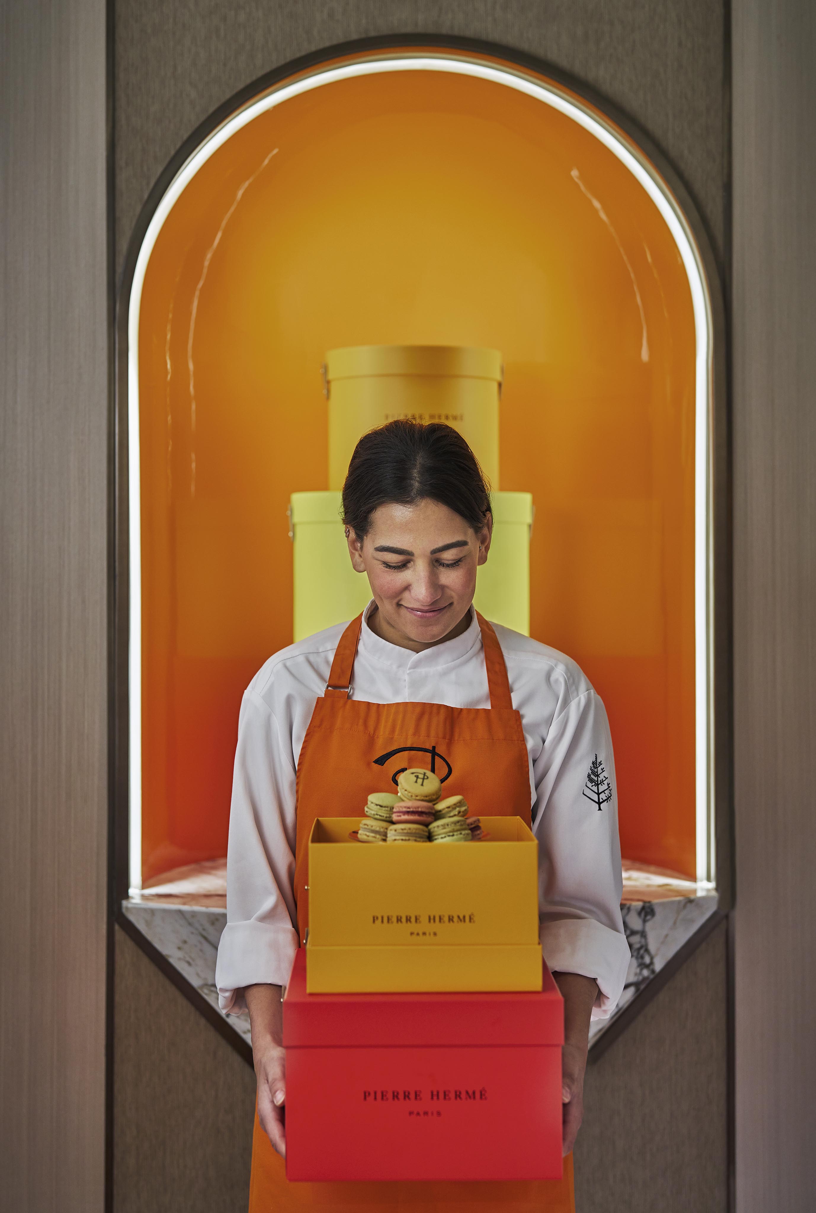 A person in a chef's uniform and orange apron holds two Pierre Hermé boxes and a plate of macarons, standing before an orange-arched display, capturing the elegance often seen in luxury hotel and resort photography in the Middle East.