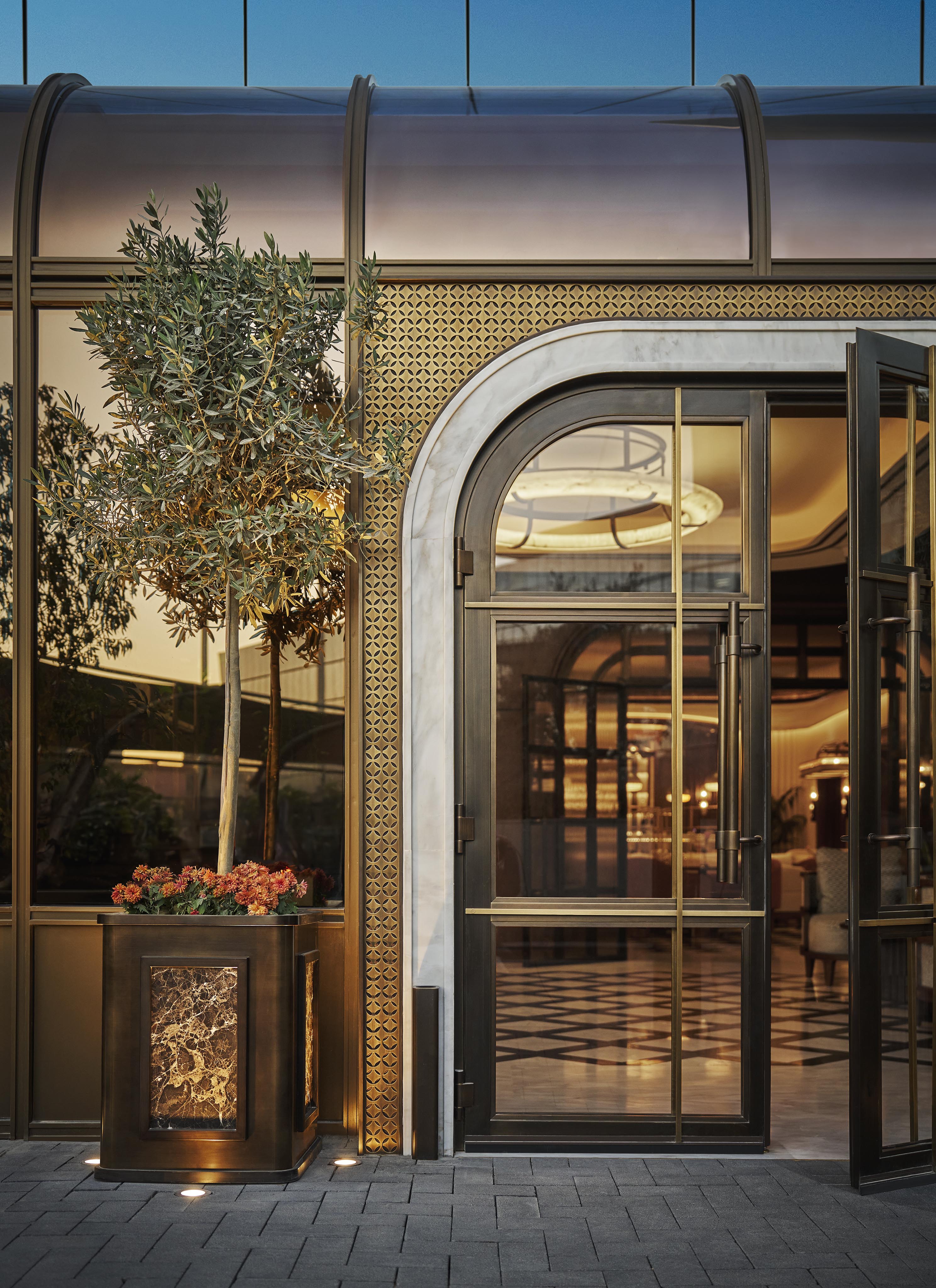 Glass and metal entrance with arched double doors, geometric decorative panels, potted plant, and warm lighting inside; checkerboard floor visible through the doorway—ideal for luxury hotel and resort photography in the Middle East.