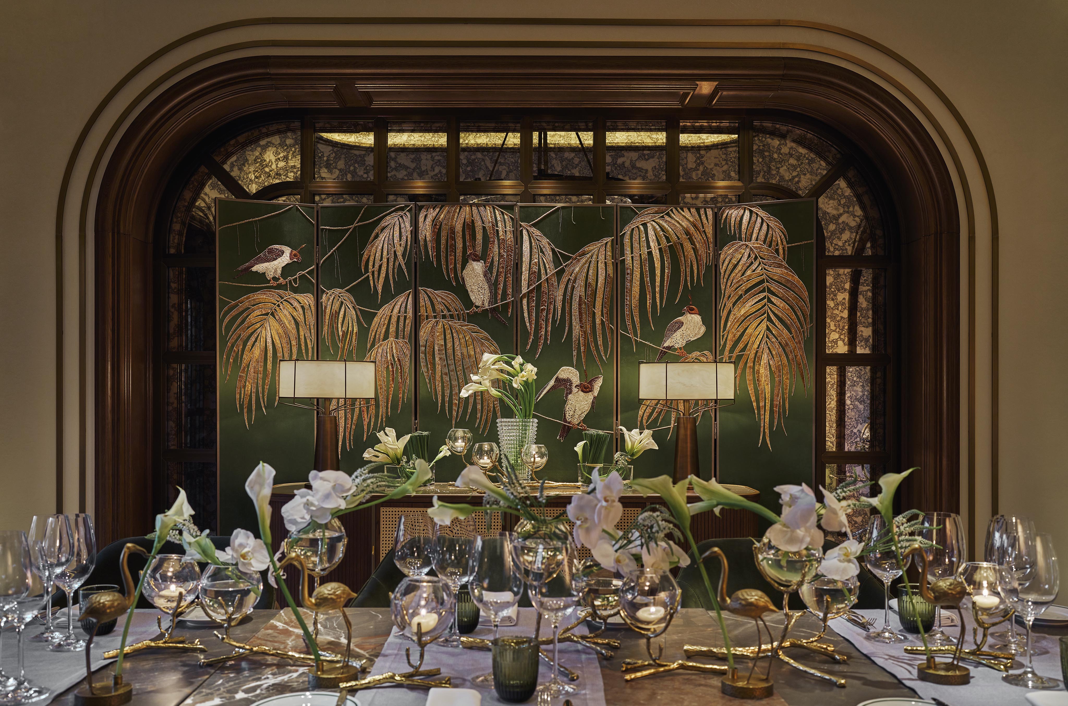 An elegant dining room in a luxury hotel, with a long table set with glassware and floral centerpieces, featuring a decorative wall with bird and leaf motifs and two table lamps—capturing the refined style of resort photography in the Middle East.