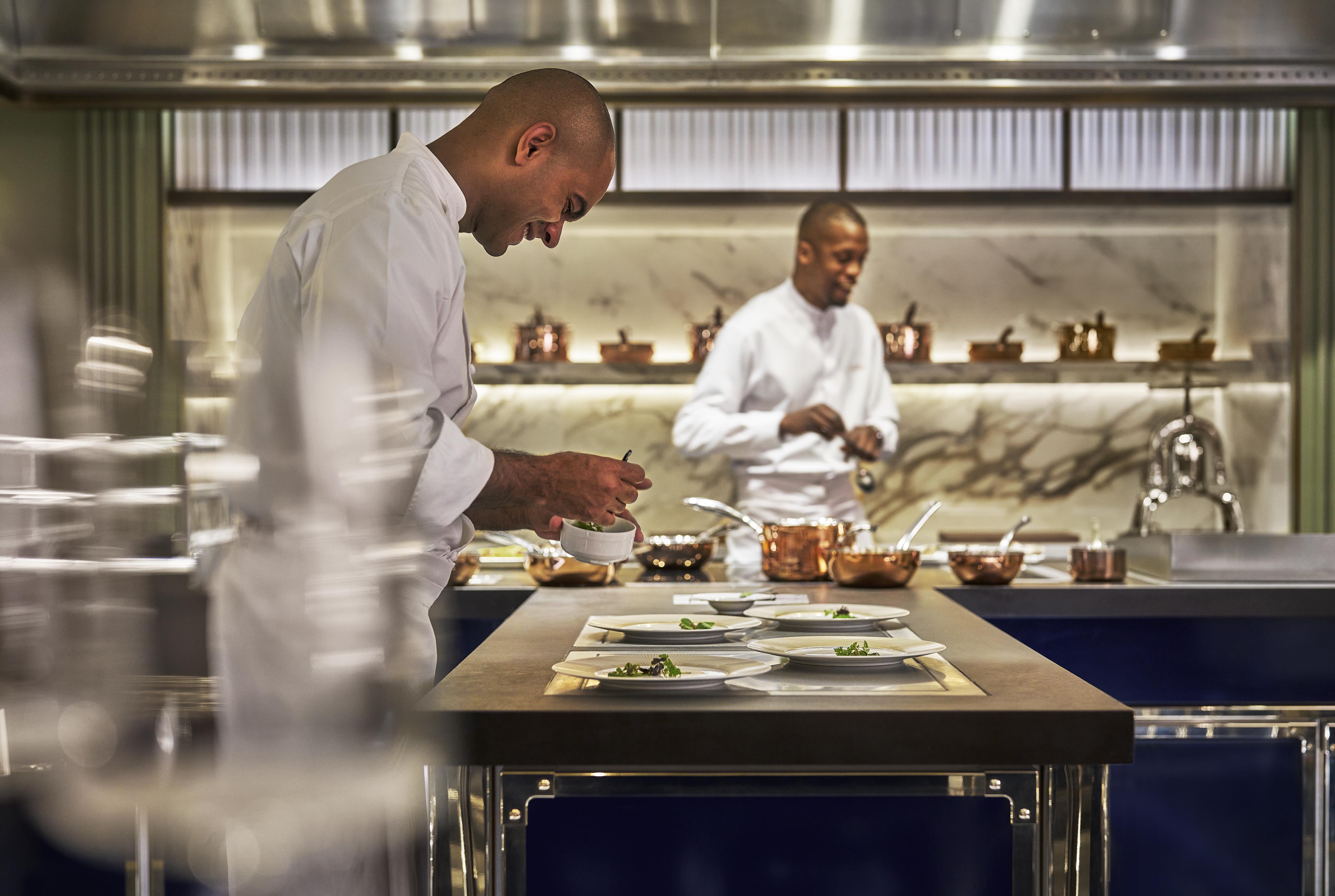 Two chefs in a modern kitchen at a luxury hotel and resort in the Middle East expertly prepare and plate dishes, focusing on their tasks with cooking equipment and fresh ingredients on the counter.